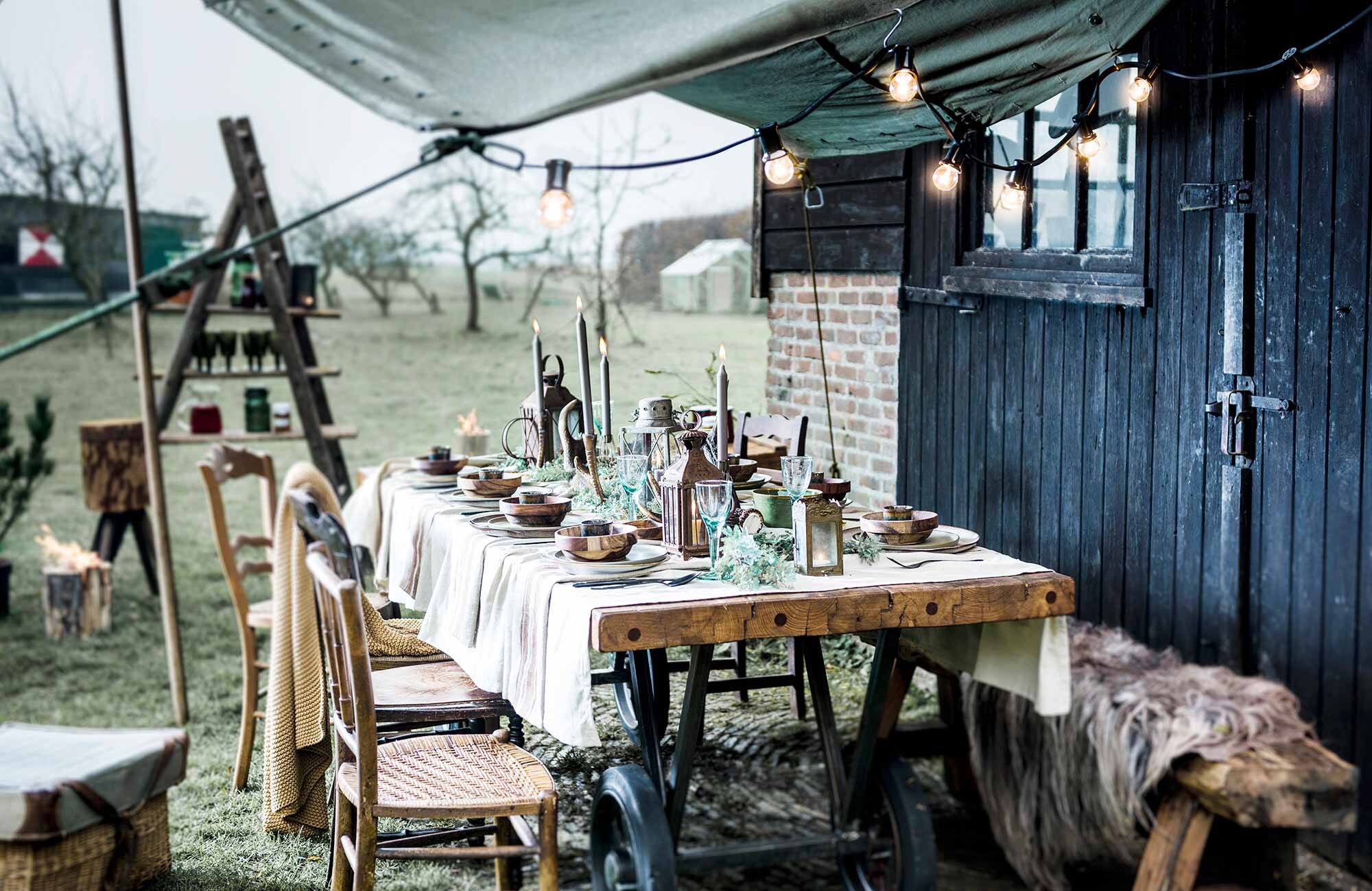 Tafel (). Bankje (). Taupe tafelkleed, okergeel plaid, wijnglazen (). Houten stoelen, groot hakblok (). Rieten mand met groene klep, ladder (). Groen legerdoek (). Bruine schapenvacht, kaarsen (). Lantaarns, grote gouden kaarsenstandaard (). Espressokopjes, houten kommetjes van HK Living, groene borden van Serax, grote wit-bruine borden van Bloomingville, vuurfakkel (). Wit bord met gouden randje (). Zwart bestek (). Bruine trui, bruine rok ()., vtwonen 03-2017 | Fotografie Sjoerd Eickmans | Styling Liza Wassenaar