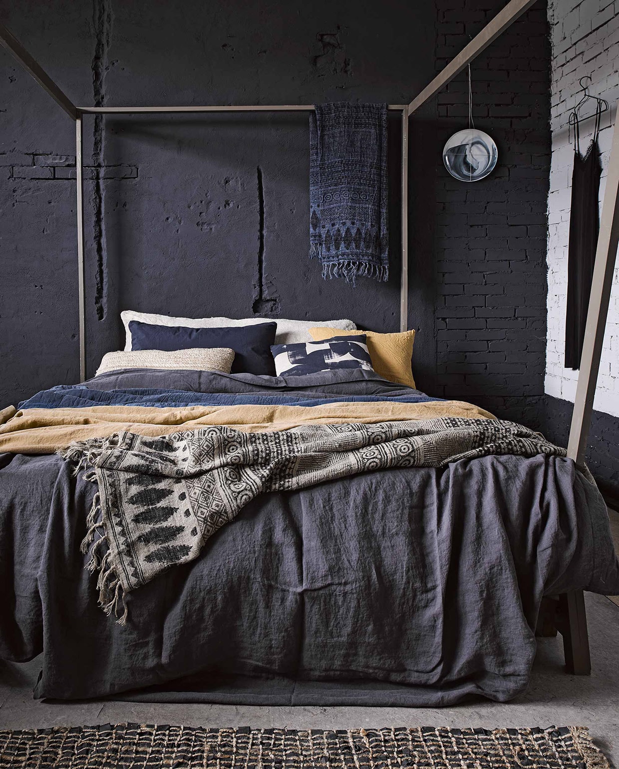 Hemelbed Safari (). Linnen dekbedovertrek in offblack antraciet, kussen met gestempeld motief (). Donkerblauwe doorgestikte sprei, linnen doek met dessin in naturel en inktblauw, vloerkleed van geweven touw met leren stroken, alles van Madam Stoltz (). Specerijengeel linnen laken van Society vanaf, donkerblauwe katoenen kussenhoes van Society (). Kussen van geweven touw (). Kerriegeel linnen kussen ). Broodzak van vlas (). Bord aan de muur (). Jurkje (). vtwonen 03-2017 | Fotografie Alexander van Berge, Hans Blomquist, Debi Treloar | Styling Marianne Luning