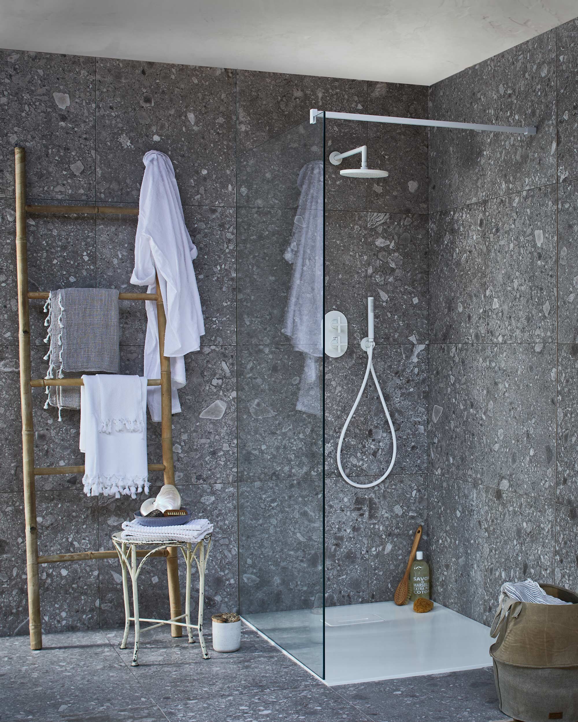 Op de wand en vloer: tegel Composite Dark Grey van vtwonen tegels by Douglas &amp; Jones, douchevloer met geïntegreerde afvoer van gegoten marmer in powder white, glazen douchewand met muurbevestiging in powder white, diverse uitvoeringen (transparant,donkergrijs en spiegelglas), douchekraan Cross, muur-versie powder white met afdekset voor inbouwthermostaat, handdouche, douchearm, hoofddouche. Bamboe ladder, witte badmat op kruk (l’Etoile Conceptstore). Hamamdoeken (Ottomania). Badborstel, nagelborstel (My Deer Store). Witte schaal (Raw Materials). Douchegel, spons (&amp;Klevering). Kruk (Neef Louis). Canvas mand (Hoffz). Schelp (De Weldaad). Pot met kurken deksel (Restored). vtwonen 02-2017 |Styling Cleo Scheulderman | Fotografie Jeroen van der Spek
