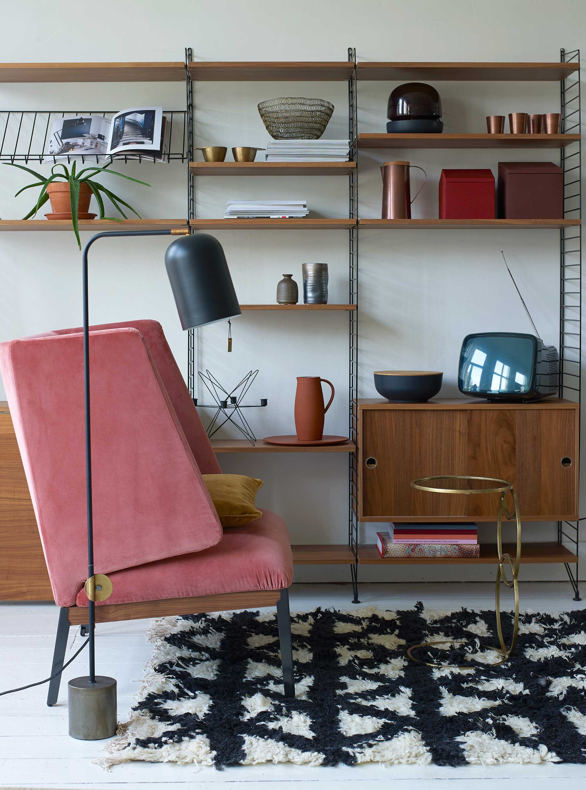 Lounge chair Hug van Arflex, zithoogte 44 cm, rughoogte 103 cm, kast String in uitvoering 200x215x30 cm (hxbxd), vloerlamp (Mobilia). Wollen vloerkleed Indochine Nights, 140×200 cm (Aai Made with Love). Bijzettafel van Madam Stoltz (deens.nl). In de kast: Kandelaar in stervorm van ijzerdraad, schaal van ijzerdraad, zwarte schaal met deksel, vaasjes van Madam Stoltz (deens.nl). Koperen kan met houten deksel, koperen bekers, messing bekers (Nijhof). Glazen lamp van Menu, terra kan en schaal van SCP, rode en bruine dozen van Menu, per set van 3 stuks (Mobilia).