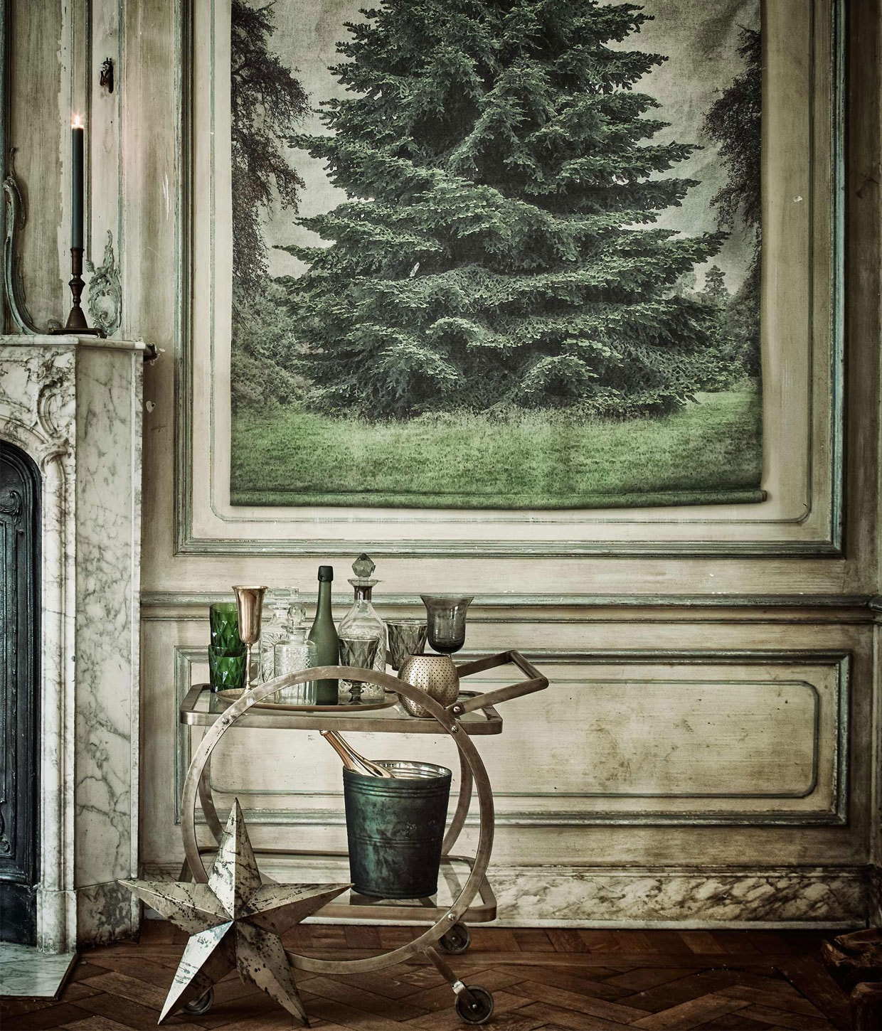 Paneel met dennenboom (). Trolley (). Groene facetgeslepen glazen, grijze glazen op voet, ronde drankfles met stop en gouden streep (). Goudkleurige ster, goudkleurig champagneglas, karaf met stop ().  van vtwonen (www.vtwonen.nl). Gouden bolvormig waxinelicht, goudkleurig ovaal dienblad (). Groene glazen pot als wijnkoeler (). Wijnflessen privébezit.