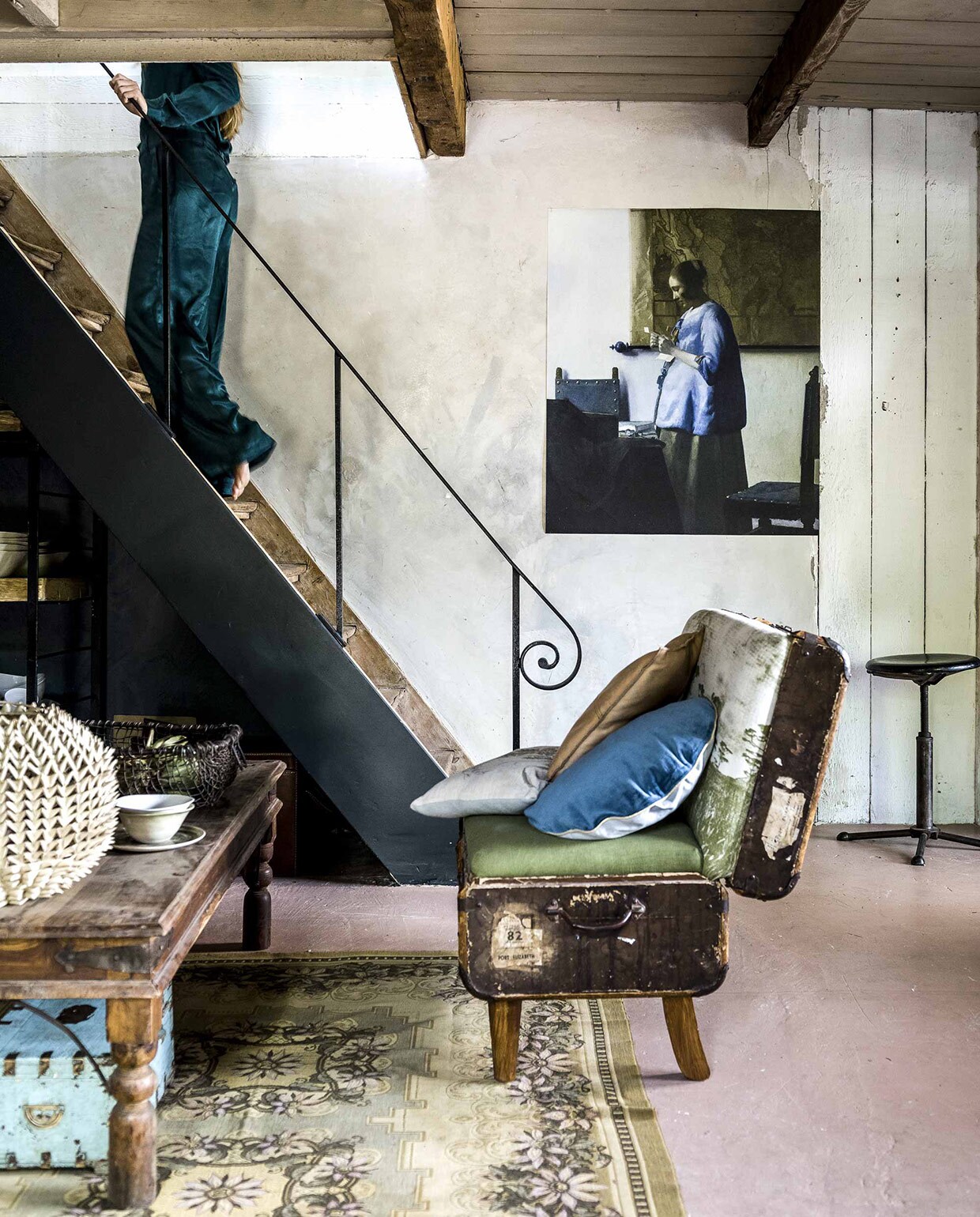 Bank van een oude hutkoffer, gemaakt in Afrika, leren kussen, kommen op tafel (),  op bank van Bloomingville, (vtwonen.nl), grijs kussen privébezit. Tafel (Jan van IJken), vloerkleed (). Lampenkap op tafel (). Mand van oude treinrails, kist onder tafel (). Kruk (), afdruk van Vermeers Brieflezende Vrouw uit de Rijksstudio op canvas (). Kleding (). Grijs kussen privébezit. vtwonen 10-2016 | Fotografie Sjoerd Eickmans | Styling Liza Wassenaar