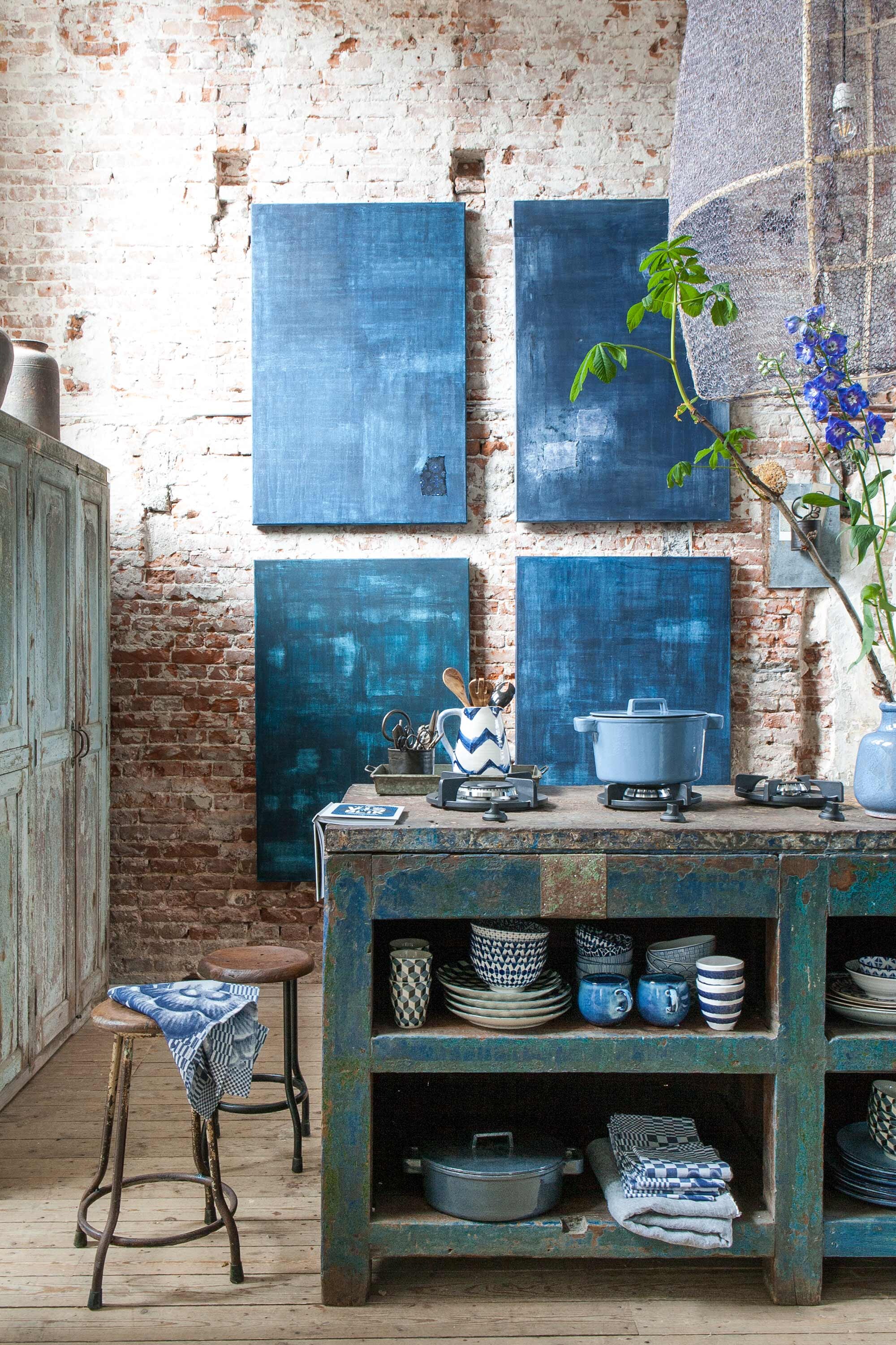Werkbank, schilderijen van Birgitta de Vos, indigo pigment op linnen canvas, kast, potten op kast, krukjes, lamp van Ay Illuminate, aluminium potje, blikken dienblad, pollepels per set van twee (Raw Materials). Industriële kookpitten prijs op aanvraag (Pitt cooking). Mokken met blokmotief per vier (Pols Potten). Pannen van gietijzer, schenkkan met zigzagmotief, vaas, theedoeken van Hollandsche Waaren per stuk, linnen doek, overige servies (Nijhof).
