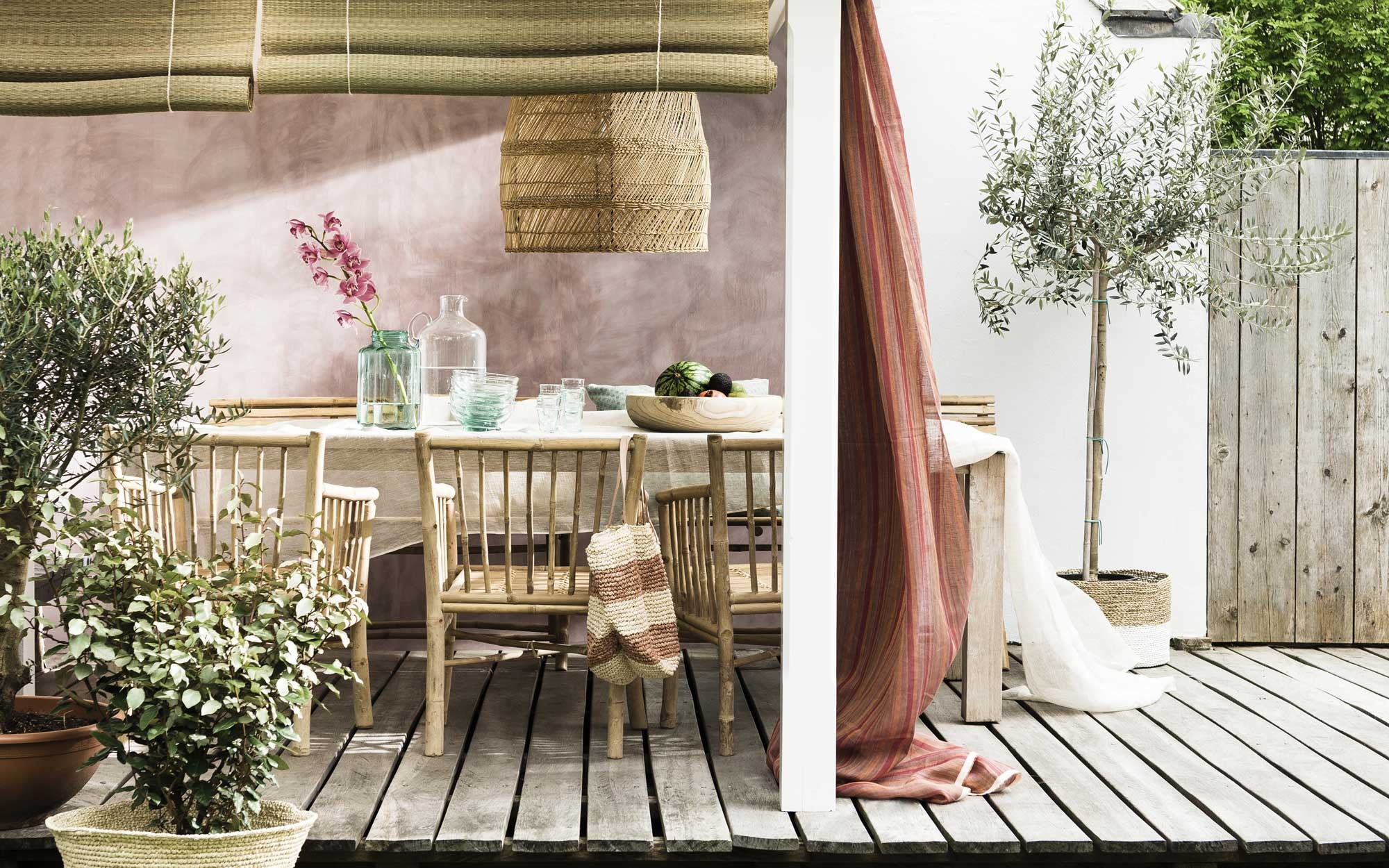Wand geverfd in kleur Dusk 24/18 van Bauwerk, muurverf (). Hanglamp l’Etoile (). Transparant linnen tafelkleed, stof (). Stoelen van Tine K Home, bamboe bankjes Mandisa van Lene Bjerre ().  Anyse Beach,  (Sissy-Boy). Grote glazen karaf, glazen schaaltjes, glazen (). Groene glazen vaas (). Mand van ananasblad, pandanmand wit-naturel (). Rieten campingmatjes, hier verwerkt tot rolgordijnen (). Gestreepte transparante linnen gordijnen(). Olijfboompjes en laurier (). Tafel privébezit.