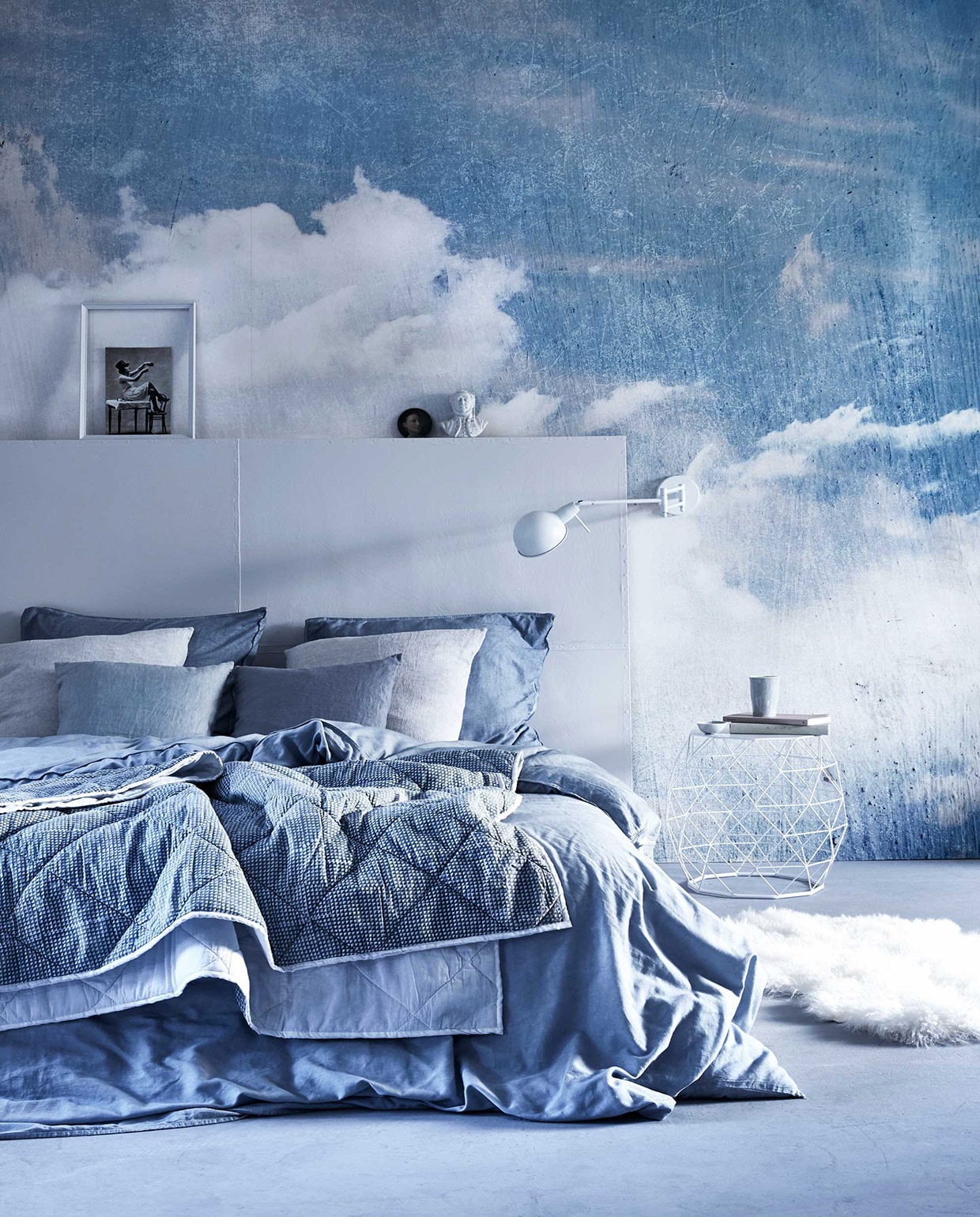 Cloud Puff van Rebel Walls (). Hoofdbord (). Beddengoed van satijnkatoen (). Beeldje (). Geprint plaid, kleine blauwe linnen kussenhoezen, schapenvacht (). Linnen  in Misty Grey (By Mölle). Bijzettafel, wandlamp Glasgow (). Onderzetter (). Fotolijst en boeken privébezit. vtwonen 05-2016 | Fotografie Alexander van Berge | Styling Cleo Scheulderman