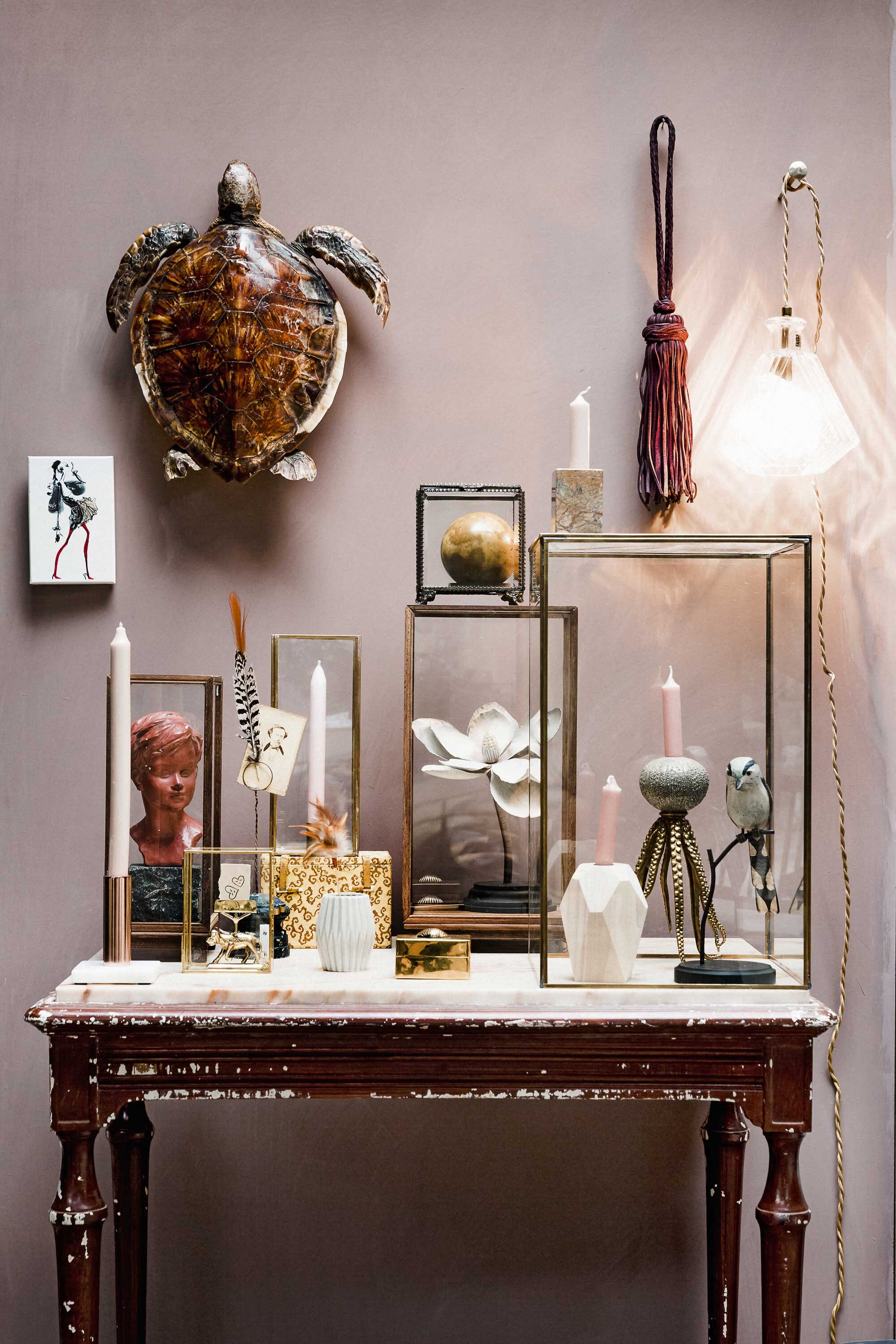 Wand geschilderd in kleur Cardamom van Bauwerk Colour (). Tafel met marmeren blad (). Lamp La Crystal van Scotch Collectables, leren gordijnkwast Le Salomé van Scotch Collectables (). Doosje met kaarten Haute Couture van Christian Lacroix van Designers Guild ().  brons met marmeren voet (Six and Sons). Stolp links (). Beeldje met hoofd (). Schuin hiervoor goudkleurige kaarthouder van Madam Stoltz met tekening van Julia Lefeber (). Papierklem (). Veertje (). Kaartje met pentekening (). Geel doosje (). Grijs vaasje (). Stolp (tweede van rechts) (). Bloem, kleine zwarte vierkante stolp (). Bol van marmer ().  met schelp (Anna + Nina). Grote stolp rechts (). Vogel van hout (). Kandelaar Octopus (). Kandelaar van licht hout ().  vierkant blokje marmer op de grote stolp, lantaarn van glas (). Kaarsen (). Schildpad aan de muur privébezit.