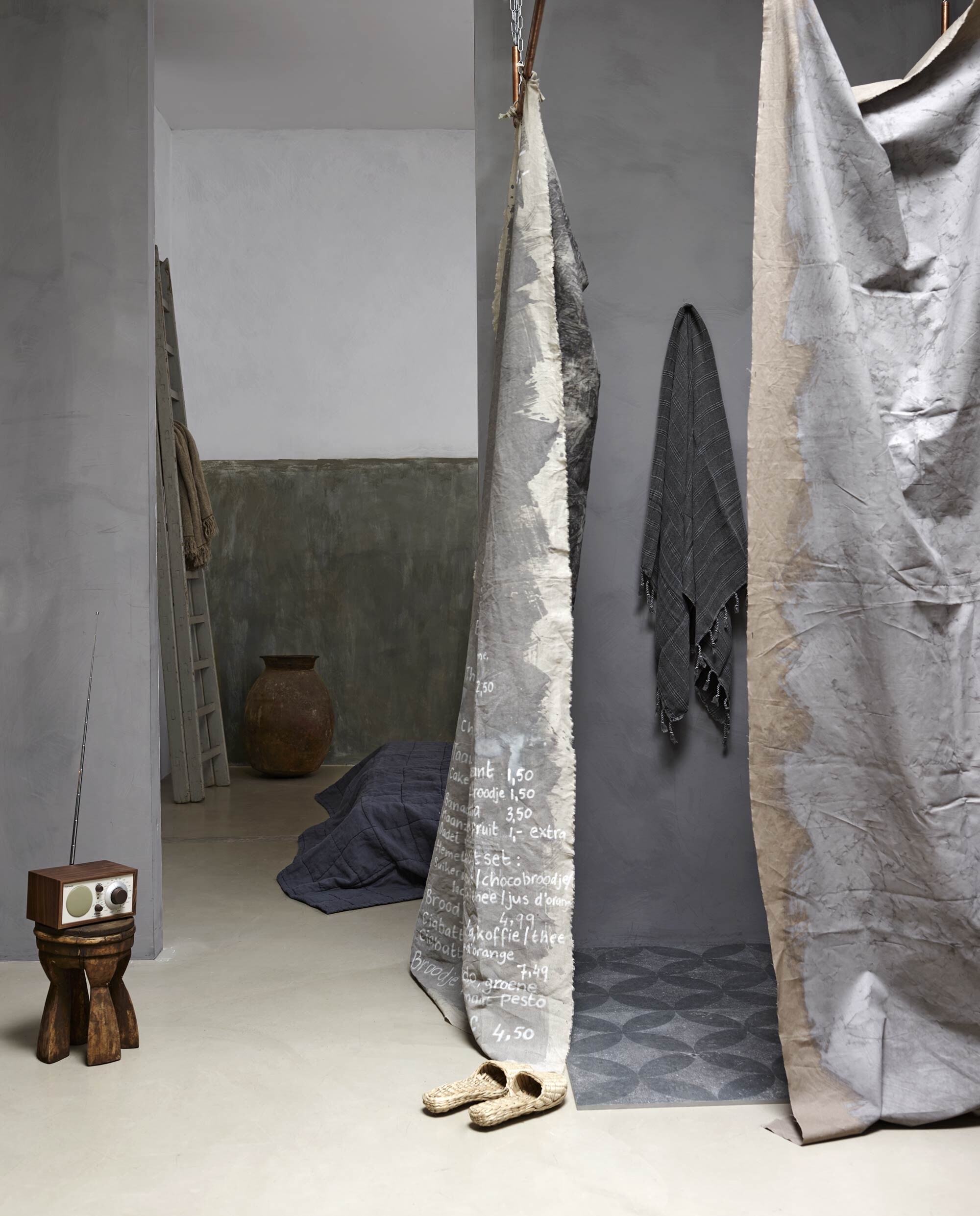 Verf Milkweed uit de Limestone collectie van Bauwerk, Frans linnen, gevlochten slippers, houten ladder, plaid van Aai made with love (AnoukB). Stone-washed hamamdoek (Hammam34). Houten krukje, aardewerken kruik (Hoffz). Tegel duostone Hormigon antracite Floret (vtwonentegels.nl). Antracietkleurige gewatteerde linnen plaid Nice (House in style).