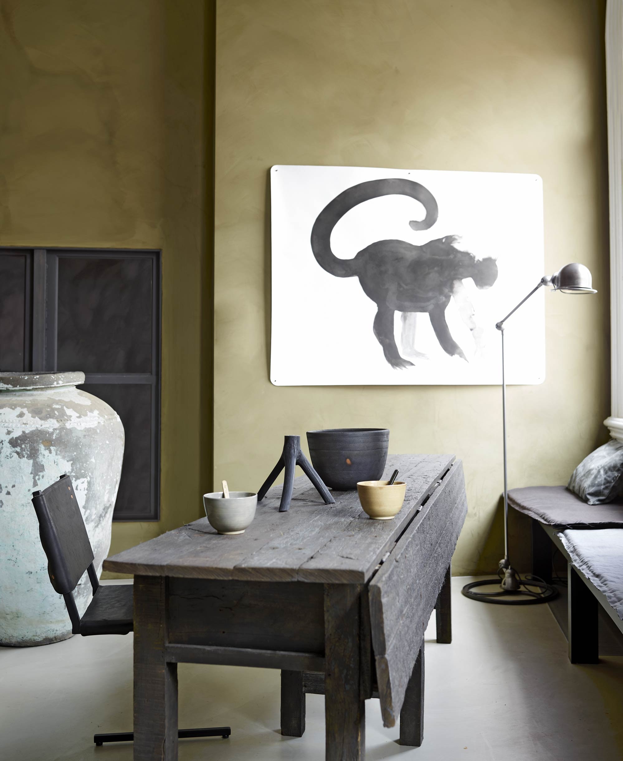 Houten tafel (Hoffz). Verf Hawtorn uit de Limestone collectie van Bauwerk, stoel van zwart leer met staal van Wdstck collective, kruik uit India, op maat gemaakte houten bank prijs op aanvraag, op maat gemaakte bank- matrasjes prijs op aanvraag, gerecycled canvas grijs kussen van Aai made with love, staande lamp Harvest &amp; Co., schaal van Muubs, kandelaar van Muubs, kom van keramiek, roerstaafje van keramiek (AnoukB). Inkttekening aap (Frank Jan van der Laan).