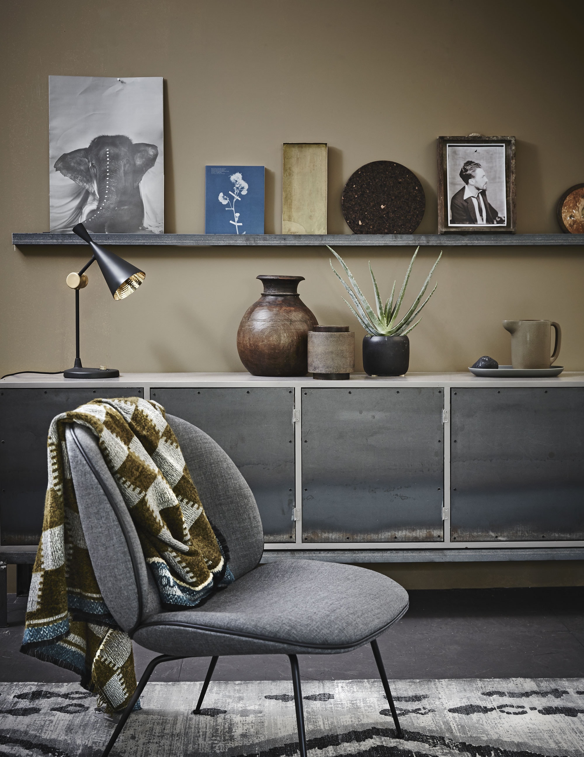 Dressoir met stalen deuren (Combitex). Fauteuil van Gubi (Mobilia). Sjaal van Humanoid (Sukha Amsterdam). Grijze kelim van HK Living (vtwonen.nl). Op de rand: papieren zak met olifant van Love Warriors (Couleur Locale), boek over blauwdruk- fotografie (Matter of Material), messing dienblad (Six and Sons), onderzetter van kurk Ø 30 cm (Ikea), laatje met foto (Six and Sons). Op het dressoir: lamp Beat van Tom Dixon (Nordic New), houten waterkruik uit India (Couleur Locale), bruine pot (Six and Sons), zwarte bloempot, kan van aardewerk, bord Sittning uit set van drie (Ikea). Muurverf Mexican Sand van Pure &amp; Original (De Ru).
