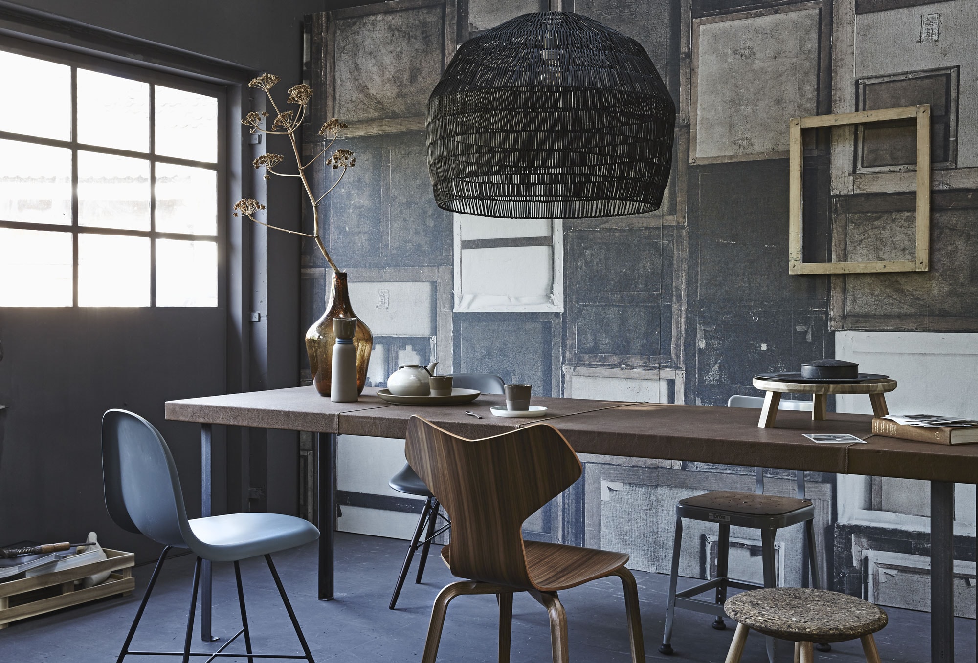 Behang Verso WDVE1501 (Wall &amp; Decò). Met leer beklede tafel (Combitex). Donkergrijze stoel links F1 van Gubi, achter de tafel een Eames-stoel (Mobilia). Midden: walnoothouten stoel Grand Prix van Fritz Hansen (Friday Next), metalen werkkruk (Spoor 38), kruk met kurkzitting (Nordic New). Vaas van House Doctor (Friday Next). Set van grijze kan en bekers (Mobilia). Ondiepe schaal, houten serveerplateau op voet (Ikea). Donkergrijs bord (Six and Sons). Marmeren doos met deksel (Hay Amsterdam). Lamp 57×72 cm (hxØ) (Couleur Locale). Houten lijst aan de wand (Spoor 38).