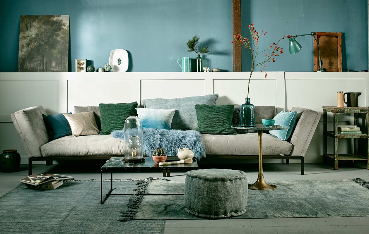 Muurverf Pure &amp; Original Classico krijtverf, kleur Libo Bay per liter, lak op lambrisering Pure &amp; Original Classico Oil Eggshell in de kleur Silver Clay, (De Ru). Long chairs City, blauw linnen kussen van Nordal (vtwonen.nl). Ecru kussenhoes van fluweel, groene gebreide hoes, blauwe fluwelen hoes met kleurverloop (H&amp;M). Groot kelimkussen (Sukha Amsterdam). Schapenvacht (Nordal). Lichtblauw kussen (De Weldaad). Vloerkleed links, ronde poef, vloerkleed rechts (Aai made with love), vaas links van bank (Raw Materials). Salontafel met groen marmeren blad en stalen frame, stolplamp (Serax). Ovaal zwart dienblad (Maison NL). Theepot (Meker Wonen). Marmeren kwast (Zenza). Hoge, ronde bijzettafel met bronzen voet (Madam Stoltz). Glazen vaas (H&amp;M). Kom (In Petto). Verweerd tafeltje rechts (Raw Materials). Kartonnen koker (Neef Louis). Kan (Ikea). Op de lambrisering, v.l.n.r. afbeelding van schilderij via het Rijksmuseum, geprint op karton vanaf (Repros), marmeren bol (Six and Sons), potje (kringloopwinkel), groene steen op voet (Fleurmonde), schaal met kever (Maison NL). Midden: Portugese kan (In Petto), groene fles (Brût Amsterdam), koraal (Fleurmonde), grijs kopje (Ferm Living). Rechts: groene atelierlamp Wielum (Brût Amsterdam), schilderdoek, omgedraaid (Six and Sons). Overig privébezit. vtwonen 11-2015 | Fotografie Jeroen van der Spek | Styling Cleo Scheulderman