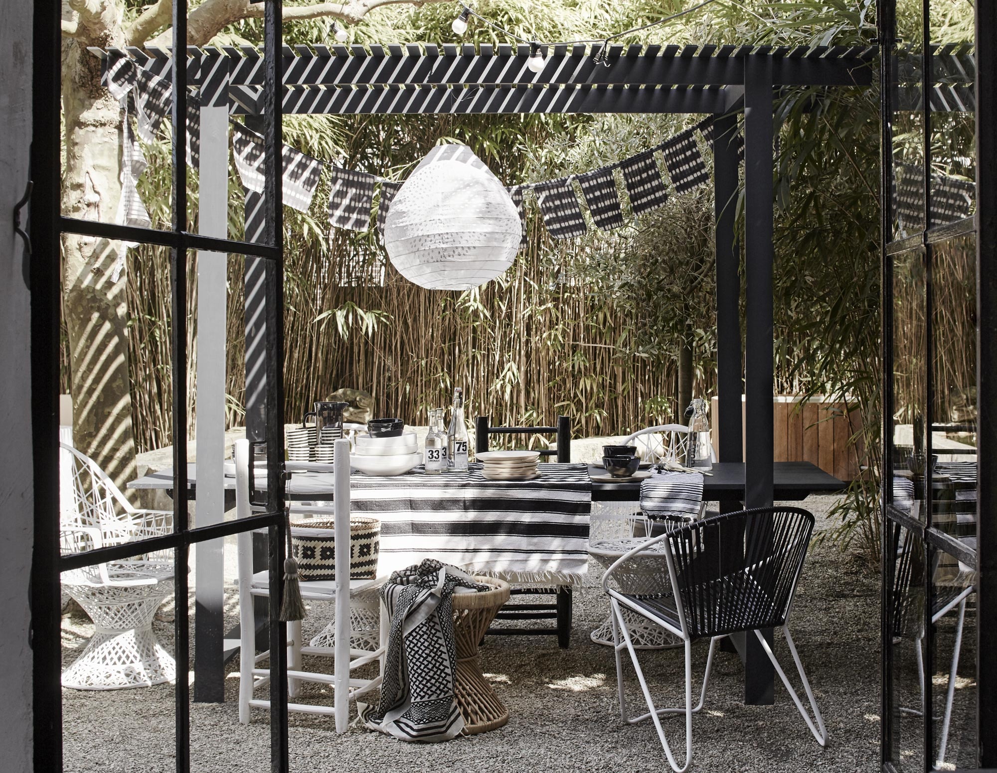 Eettafel (Combitex). Stoelen naast en achter de tafel: witte gevlochten buitenstoelen (Household Hardware). Zwarte Marokkaanse stoel van HK Living (vtwonen.nl). Stoelen voor: witte Marokkaanse stoel (vtwonen.nl). Rotan kruk van Tine K Home (l’Etoile Conceptstore). Tuinstoel met wit frame en zwarte zitting van Nordal (vtwonen.nl). Stoffen lampion Micha Gold Drop, Ø 50 cm (It’s Loft). Zwart gestreept tafelkleed en servet (www.nooosugar.nl). Houten lepels, gestreepte mokken, witte en zwarte schaaltjes, witte diepe borden (Household Hardware). Shawl (&amp;Klevering). Rieten mand op stoel (Tierlantijn). Houten borden van HK Living (vtwonen.nl). Limonadeflessen (Tierlantijn). Kunststof kan (H&amp;M).