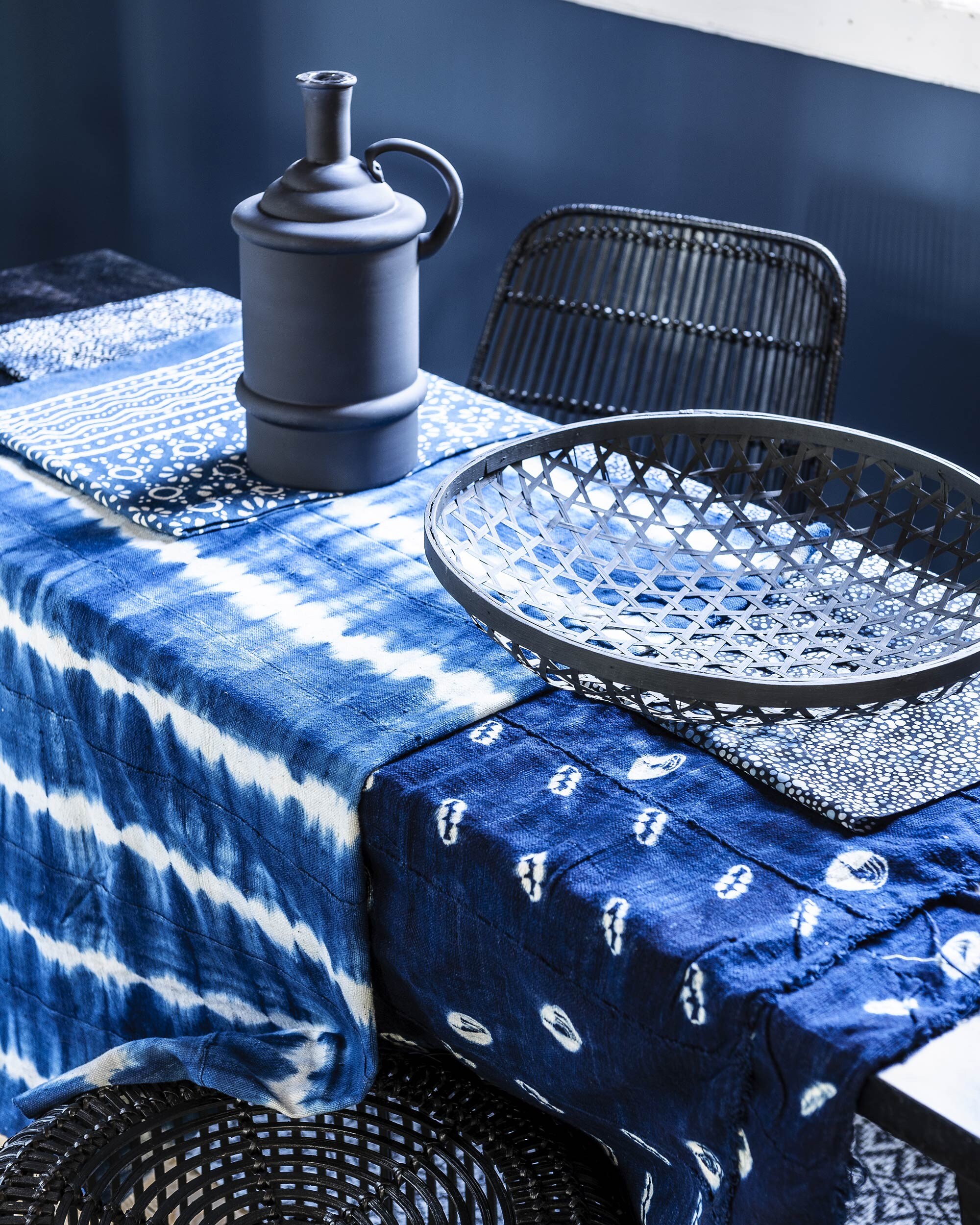Eetkamerstoel van zwart rotan (). Zwarte bamboe kruk ().  van Madam Stoltz (vtwonen.nl). Zwarte mand (). Batik tafel- kleed met bloemdessin 150×220 cm (). Batik doek met stipjes van Hoffman (). Eco-katoenen indigo doeken 115×150 cm ().  Stack van House Doctor, 90×200 cm (vtwonen.nl). Wand geverfd met extra matte muurverf in de kleur Deep Steel ().