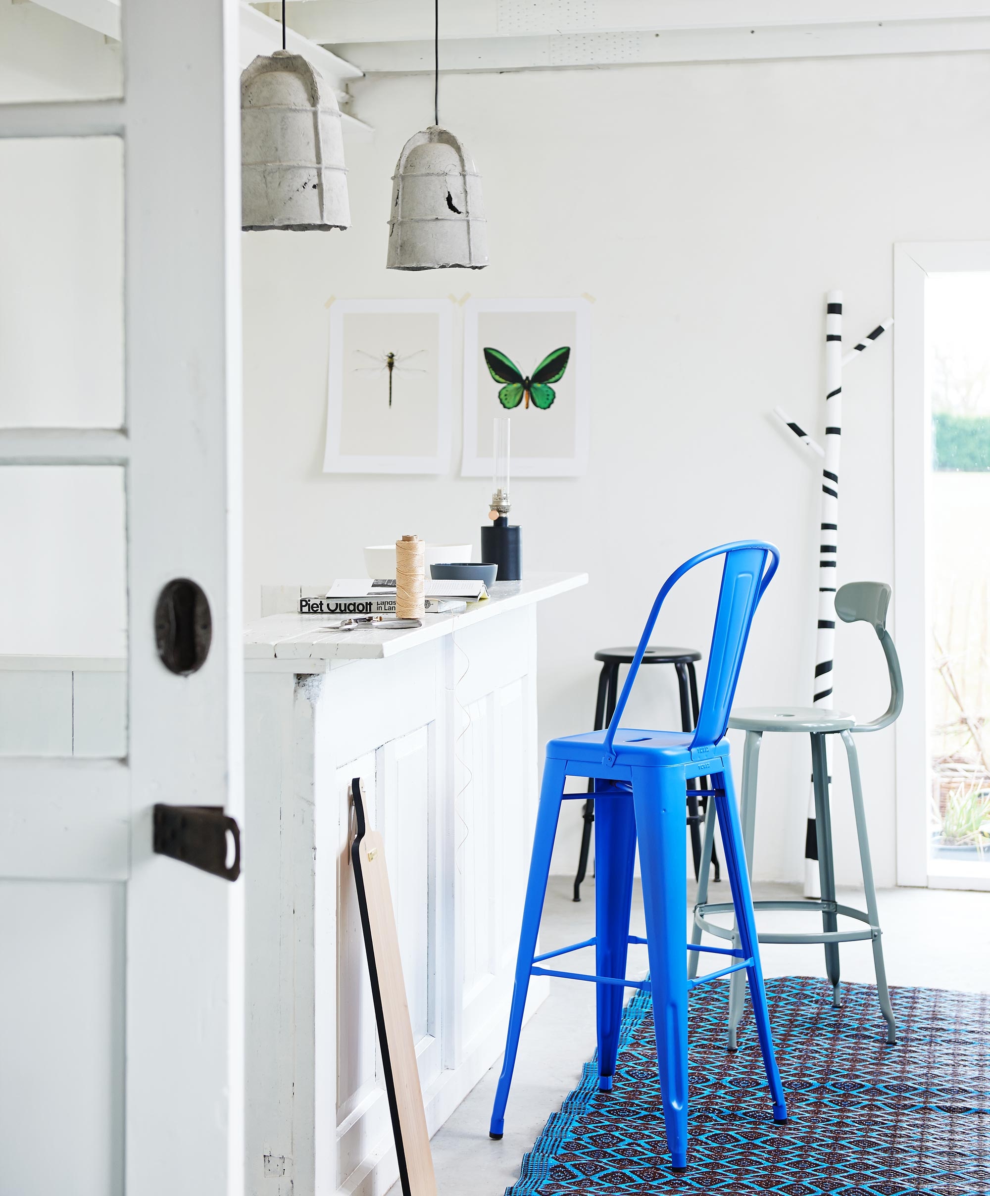 Hanglampen Mold van Eternit, blauwe kruk Grand Dossier van Tolix, groene barkruk van Chaises Nicolle met rugleuning, 75 cm hoog, zwarte barkruk zonder leuning van Chaises Nicolle, 75 cm hoog, snoeischaar van Sophie Conran (Extra Vert). Poster van vlinder en libel, broodplank (Nijhof). Olielamp Patina (Urbans and Indians). Grote witte kom Bamboo, kleine grijze kom Bamboo (Hema). Houten stok in wit met zwarte strepen (HK Living). Kunststof vloerkleed 120×200 cm (Urbans and Indians). In de potten en balkonbak zit Rozemarijn.