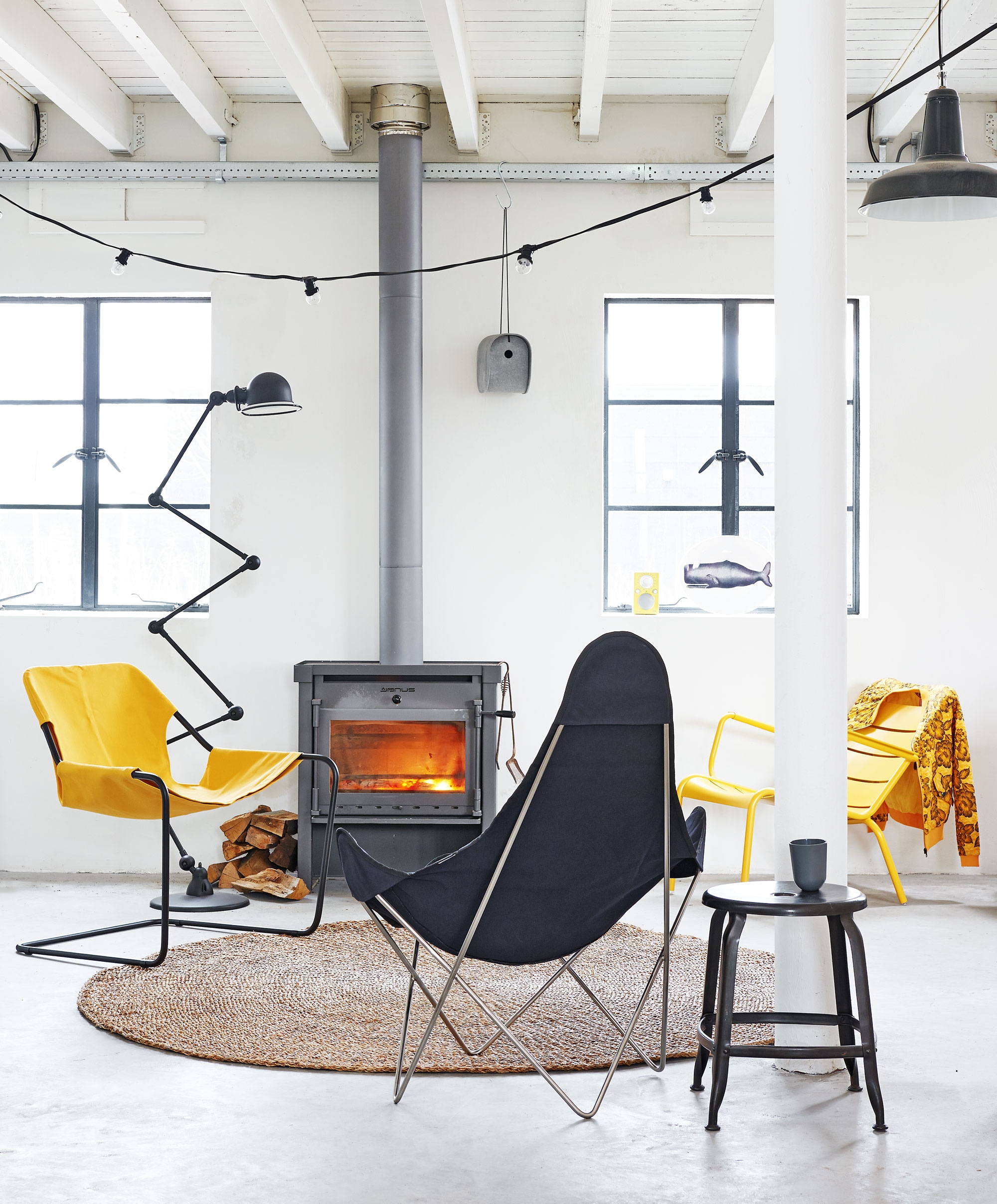 Geel-zwarte stoel Paulistano Chair met katoenen hoes, zwarte stoel Beatle chair met katoenen hoes, gele stoel Luxembourg low armchair van Fermob, zwarte kruk van Chaises Nicolle, vogelhuisje Birdy van Eternit (Extra Vert). Vloerkleed van zeegras Ø 200 cm, Tivoli Audio radio Pal (niet meer leverbaar in geel) (Nijhof). Jieldé vloerlamp (themanor.nl). Melamine mok (Hema). Snoerlamp light chain van House Doctor (vtwonen.nl). Schaal van melamine met walvis (Urbans and Indians). Jasje (Edith &amp; Ella).
