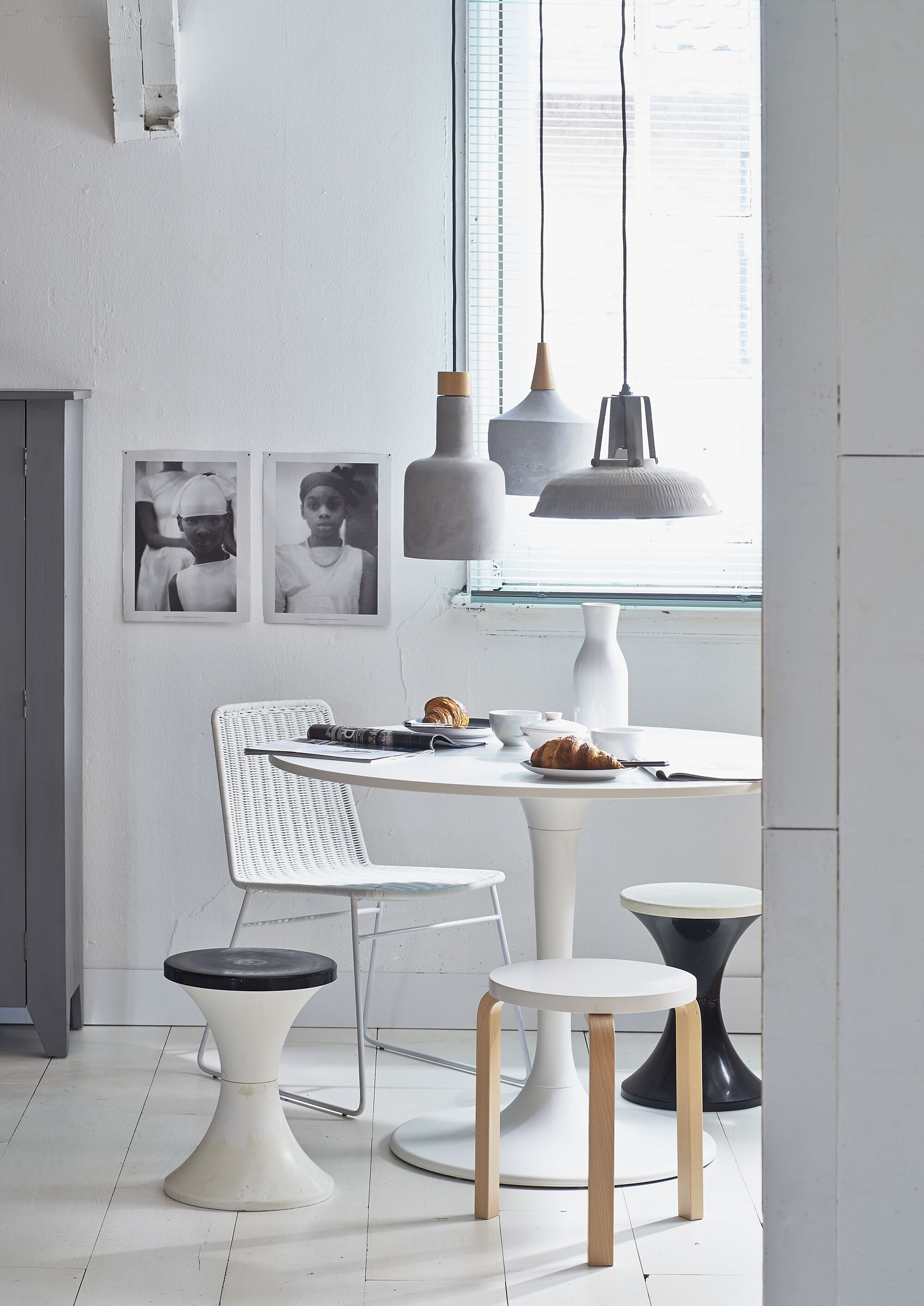 Tafel Docksta Ø 105 cm (Ikea). Witte rotan stoel, betonnen lampen Carafe en Bottle van Dutchbone per stuk (DR Wonen). Soortgelijke houten kruk met witte zitting Frosta (Ikea). Kunststof diabolo-vormige krukjes, witte aardewerken karaf, kommen per stuk (Van Dijk &amp; Ko). Borden per stuk (Ikea). Metalen lamp (De Weldaad). Fotolijsten privébezit.