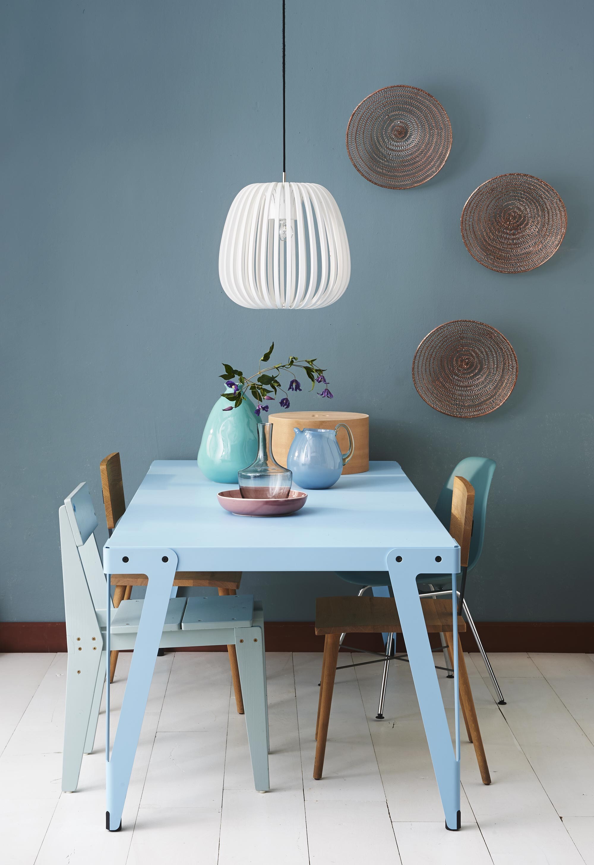 Metalen tafel Lloyd 200×90 cm (Anno). Houten stoelen Dowel, aan de wand manden Copper per stuk (DR Wonen). Plankstoel (Floris Hovers). Eames-stoel in ocean blue (Vitra). Turkooise vaas (De Weldaad). Roze schaal (Nijhof). Karaf Hämta (Ikea). Blauwe glazen kan Susa van Casa Vivante, lamp Bamboo van Ay Illuminate (vtwonen.nl). Houten doos Hat (Hay).