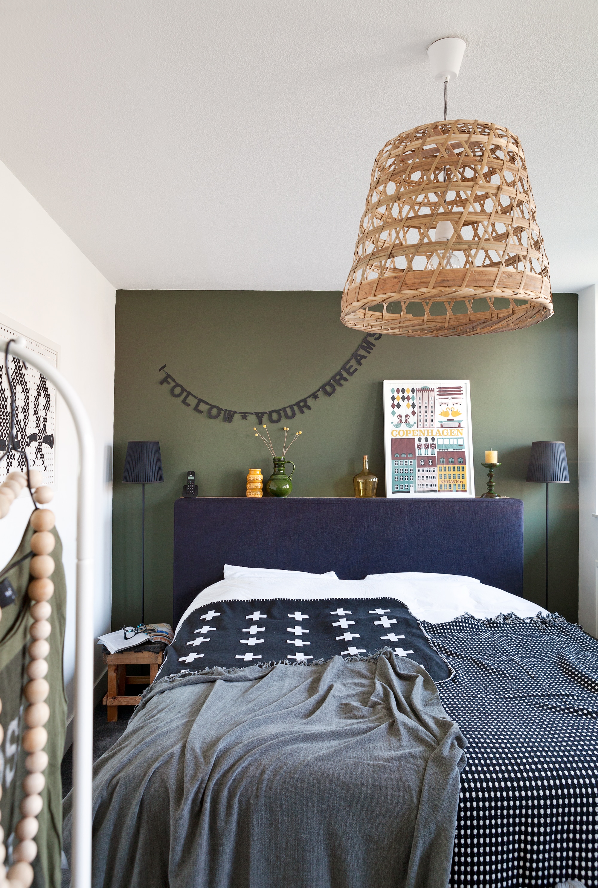 De slinger met zelfbedachte tekst is gemaakt van losse letters, Make your own Beamer, te koop bij . Marc maakte een steigerhouten frame en plaatste dat achter het hoofdbord van het bed, zodat Lisanne daar accessoires op kan zetten. De ingelijste theedoek van Ferm Living, een geel en groen vaasje van de kringloopwinkel, een glazen vaasje van HK Living en kandelaar van Hema. Lampjes van Ikea, krukje met zitting van oude banden van Intratuin. De deken met de witte plusjes is door Lisanne zelf gemaakt, de zwart-witte en grijze plaid komen van Ikea. De ketting is zelfgemaakt, net als de lamp, ooit een vtwonen zelfmaakidee, gemaakt van een mand die is gekocht bij de Fair Trade winkel.