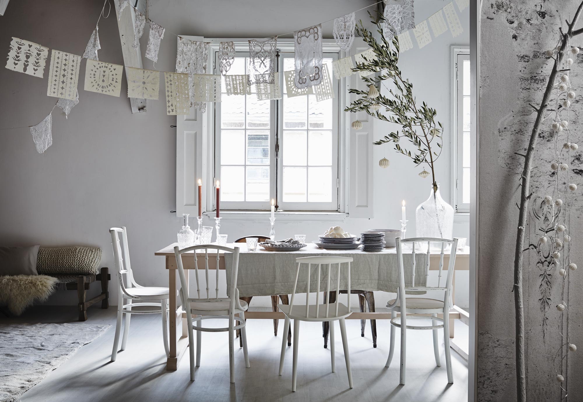 Tafel (Combitex). Oude spijlenstoelen, glazen vaas (De Weldaad). Moderne spijlenstoelen van Hay (Mobilia). Metalen stoel Tolix (Neef Louis). Linnen laken als tafelkleed 250×290 cm van La Fabbrica del Lino (Fich Home). Servies (Pottenbakkerij Anouk). Bestek, kanten kleedjes gebruikt als slinger (Jolandelijk). Glazen met stip, glazen kristal, glazen karaf, glazen kandelaars, witte papieren kerstdecoratie, lichtsnoer aan tak (Nijhof). Kaarsen (Hema). Daybed, slinger van geknipt papier, gebreid kussen, schapenvacht, vloerkleed van Private (Sukha). Kussen silver, coated linnen 45×60 cm (Mrs. Me). Wall &amp; Decò behang Stucco (De Ru).