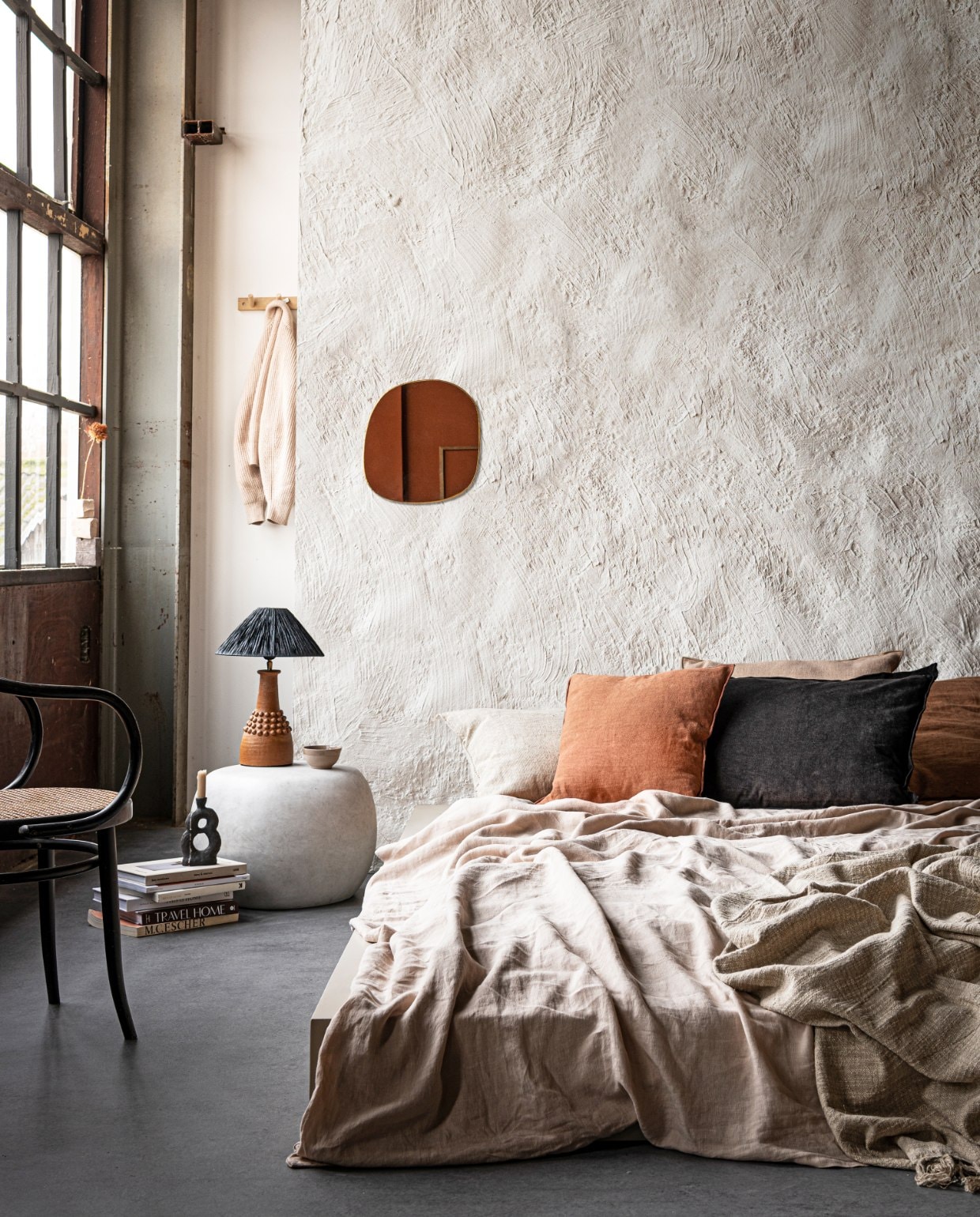 Muurverf in Chalk White (pure-original.nl). Bed op maat gemaakt en geverfd met Farrow &amp; Ball in Oxford Stone (com-anders.nl). Linnen dekbedovertrek in Sand van Linge Particulier, vanaf € 325,-, kussen Sabra offwhite Oblong XL € 113,-, linnen kussen Vice Versa van Maison de Vacances € 133,-, kussen Vice Versa Canvas Formentera van Maison de Vacances € 207,-, kussen Vice Versa Toile Nomade van Maison de Vacances € 161,-, messing spiegel Ame van Caravane € 141,- (couleurlocale.eu). Zwart kussen van vtwonen collectie € 34,95 (vtwonen.nl). Bijzettafel Pebble van vtwonen collectie € 129,-(basiclabel.nl). Lamp van Madam Stoltz € 231,75 (deens.nl). Kandelaar van Malwina Kleparska € 95,-, boek van Bea Mombaers € 43,50 (studio-yen.com). Kom van klei € 9,- (household-­­ hardware.nl). Kapstok van ByOn € 24,95 (littledreamers.nl). Stoel en overige boeken privébezit. vtwonen 03 2021 | styling Marlies Does, fotografie Sjoerd Eickmans