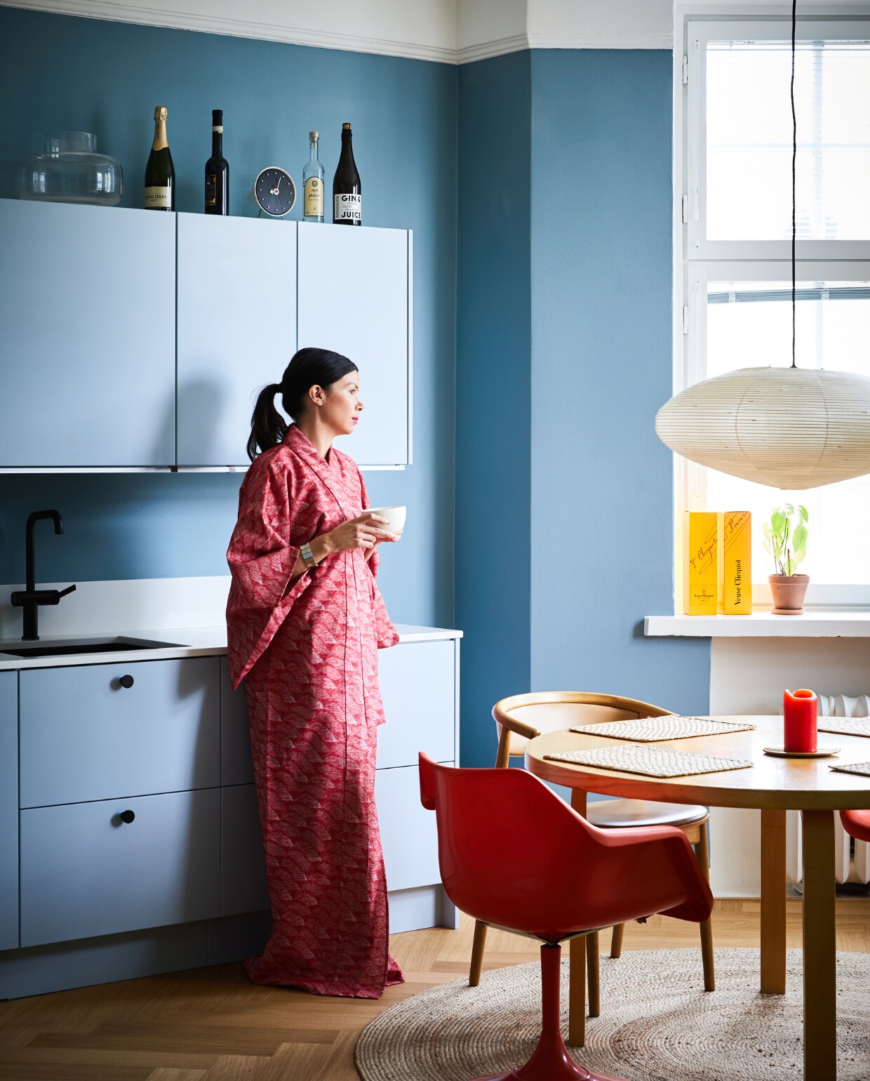 Ook de keuken kreeg een lichtblauwe tint. Eettafel en placemats Artek. Vintage oranje stoel Robin Day voor Hille, houten stoel Mama Hakola. Klok en lamp Akari Vitra. Vloerkleed Anno. NEE | vtwonen binnenkijken special 12-2022 | Productie KJonna Kivilahti | Fotografie Krista Keltanen/Living Inside | Tekstbewerking Rosa Suikers