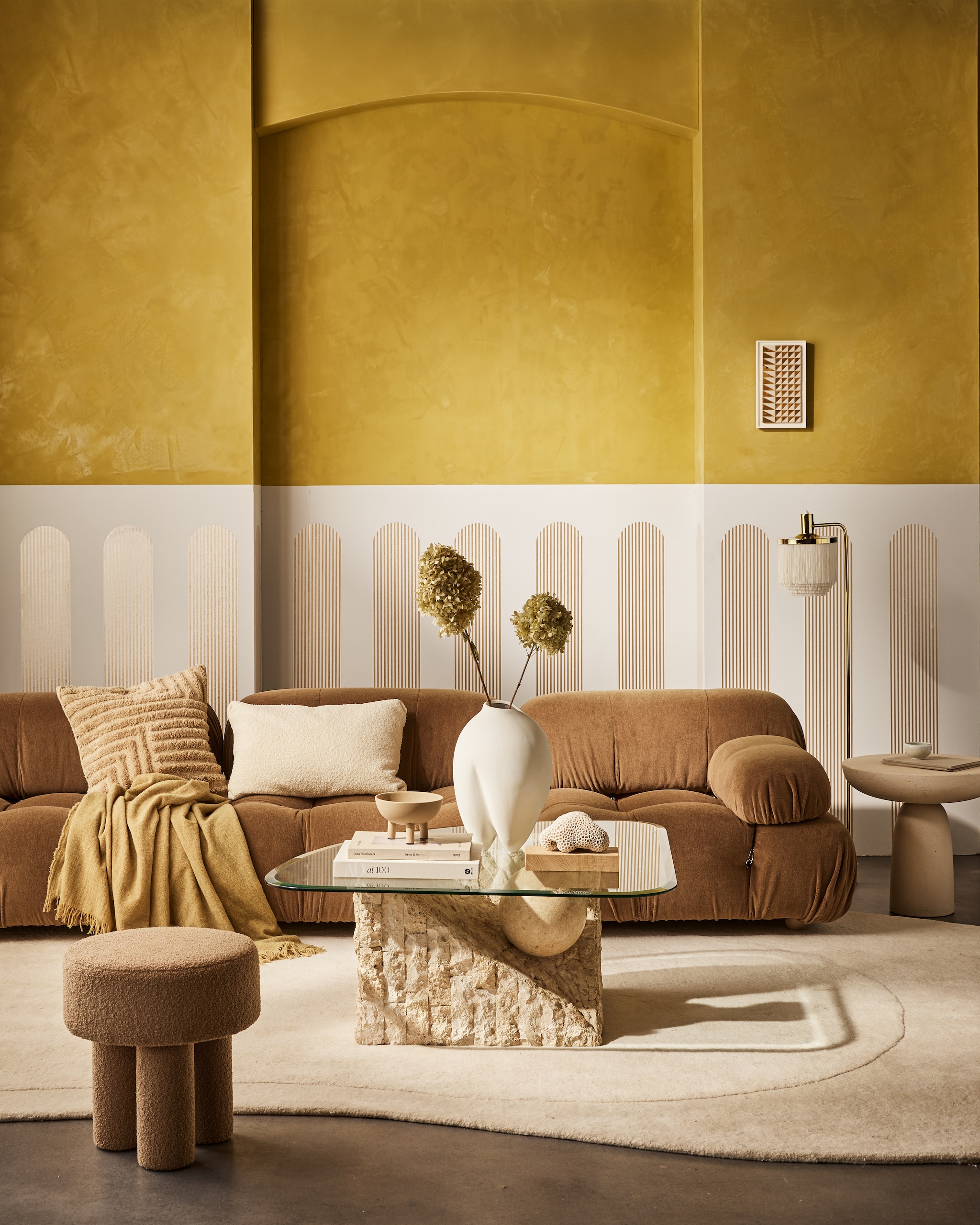 vtwonen collectie behangstrook Streep Gold-Egg White, 17,6 cm (b), rol van 10 m. M.m.v. homestede.nl (bank, kussens, plaid), edha-interieur.nl (poef, bijzettafel), goeds.nl (vloerkleed van Layered, lamp), pietjonker.nl (salontafel), rollmannceramics.com (kunsttegel), lauthentique.nl (gele muurverf kalkverf in Korengoud 33, witte muurverf krijtverf in Krijt 63), 101cph.com (vaas, schaal). Overig privébezit. vtwonen collectie 02-2023 | productie Tanja Saarberg | styling Cleo Scheulderman | fotografie Jeroen van der Spek | assistentie Kelly van Gendt | studio en decorbouw Studio Milo