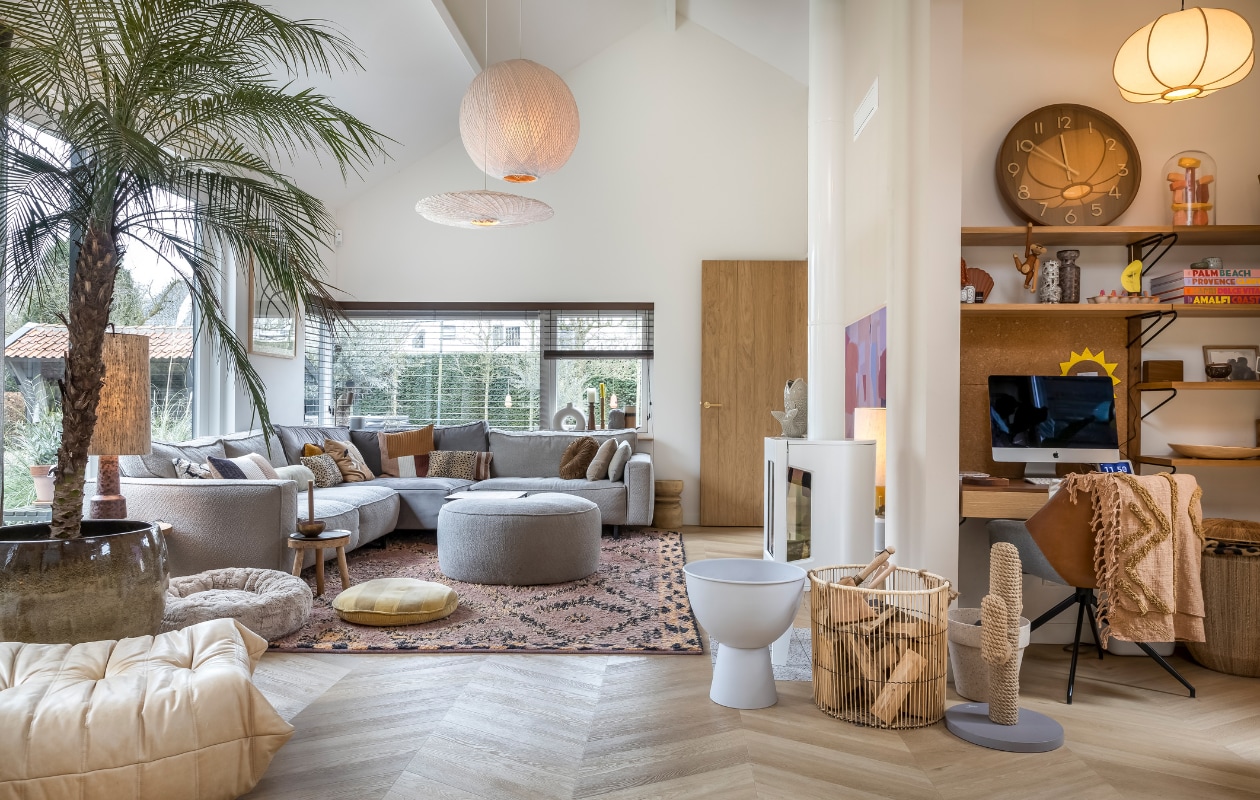 Muurverf Mushroom van Little Greene, verf plafond RAL 9010, pvc-vloer Therdex Chevron 6541. Hanglamp rond en ovaal HKliving. Kandelaars in vensterbank van links naar rechts: HKliving, Loft, grijze vaas onbekend, HKliving. Bank Melchior Interieur, kussens op bank: Ferm Living en HKliving. Kruk rechts van bank Karwei. Hocker grijs Melchior Interieur. Vierkant tafeltje, rond tafeltje, vloerkleed en geel kussen HKliving. Hondenmand dierenwinkel. Lamp op tafeltje HKliving. Plantenpot Jasmijn Bloembinders, palmboom Green Bubble. Togo fauteuil Ligne Roset. TV Samsung The Frame. Houtkachel Jotul, vis op kachel H&M, lamp achter kachel en witte plantenbak HKliving. Mand Karwei. Krabcactus Designed by Lotte. Bloempot Karwei. Wandkast Made (niet meer verkrijgbaar). Klok Karlsson. Stenen onder stolp interieurwinkel in Los Angeles. Aapje Kay Bojesen. Vaasjes HKliving. Stoel Zuiver, plaid op stoel HKliving. Mand rechts van stoel Loods 5. Hanglamp H&M. Raambekleding Onel Window Dressings. Overig onbekend. vtwonen 02-2023 | styling Karin van Schie | fotografie Henny van Belkom