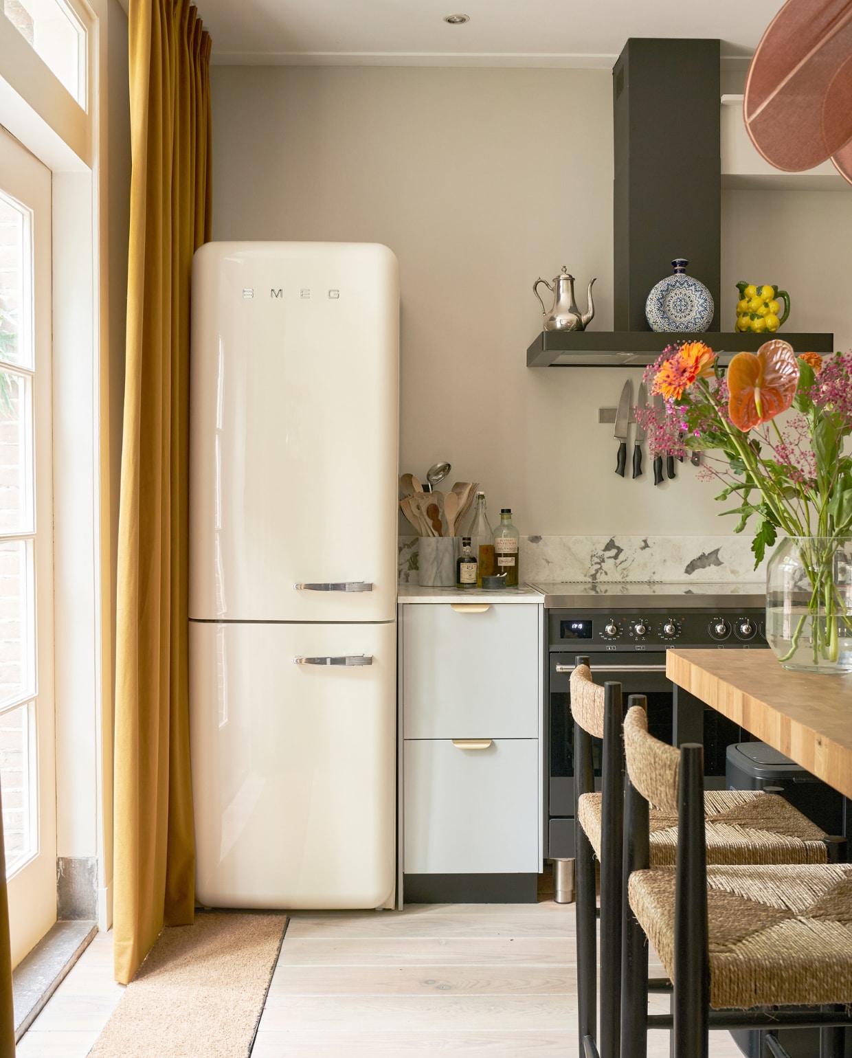 Fornuis en koelkast Smeg, marmeren pot meegenomen uit New York, blauwe karaf uit Mexico, zilveren theepot van Emilie’s grootmoeder, citroenenkaraf &amp;klevering, gordijnen Veneta, barkrukken Zara Home, keuken Veddinge en keukeneiland Ikea, marmeren blad Jan Reek Natuursteen, vaas Bloomon, messing greepjes Luste van Meraki, keukengerei en messenrek onbekend. vtwonen 02-2023 | productie CocoFeatures.com | fotografie Muk van Lil