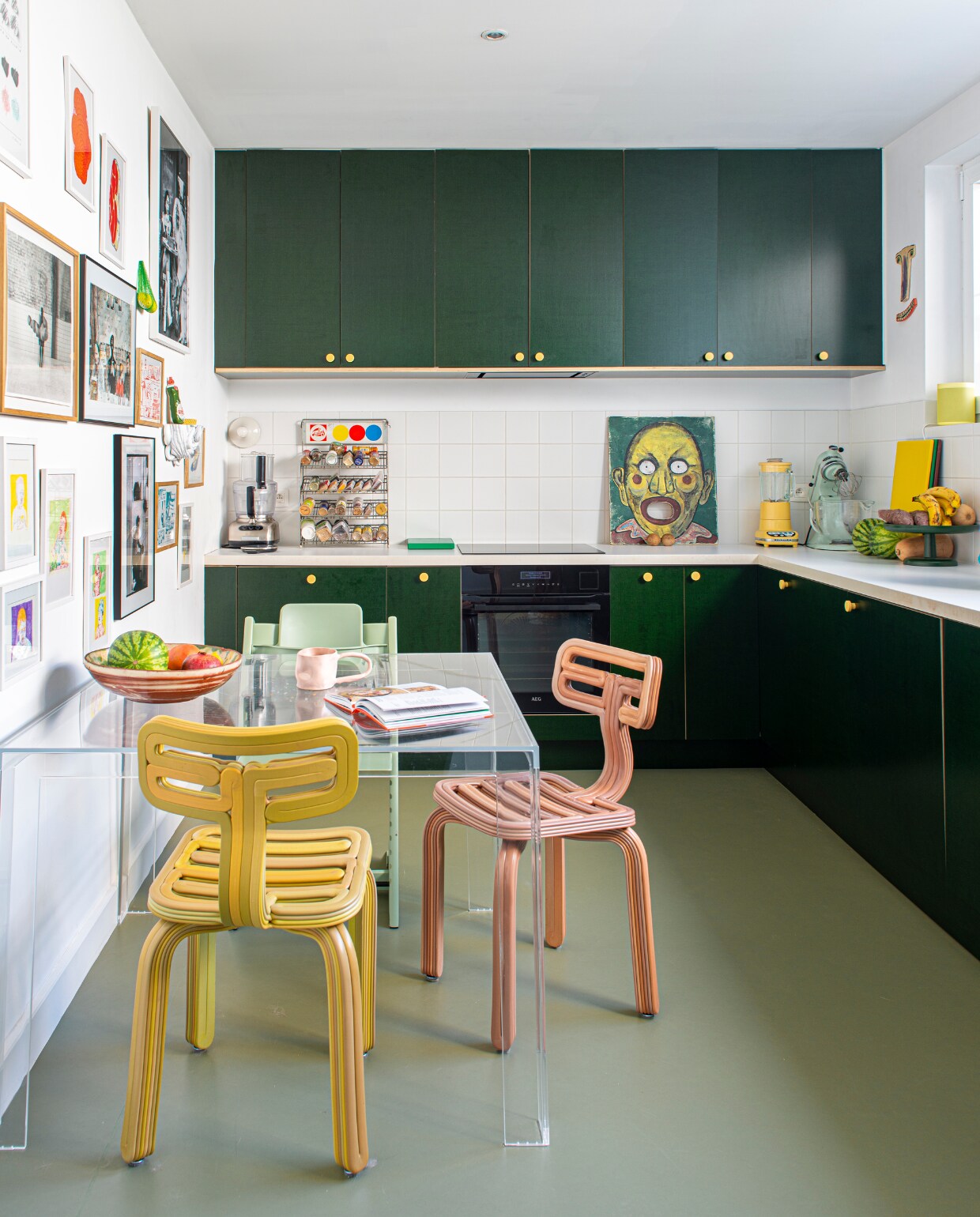 Keukenkastdeuren zelfgemaakt van betonplex met handgrepen van Toniton. Vloer Marmoleum in Rosemary Green van Forbo. Stoelen Chubby van Dirk van der Kooij, tafel Invisible van Tokujin Yoshioka voor Kartell. Blauw object Mommy I made an Ashtray van Rutger de Regt, fruitschaal Vitra, gele speakers Hay x Sonos, naast raam keramisch gezicht Choses Faites Mains, schilderij op aanrecht vintage, spel passe-boules, oud verfdisplayrek van Talens gebruikt voor kruiden. Wandlamp Teti van Artemide. Aan de wand kunstwerken van onder anderen Louise Daneels, Mickey Philips, Hélène Jean-Bon, Joost Stokhof en Mark Bohle. Overig onbekend. vtwonen 02-2023 | productie De Binnenkijkers | styling Jeske Weel | fotografie Caroline Coehorst