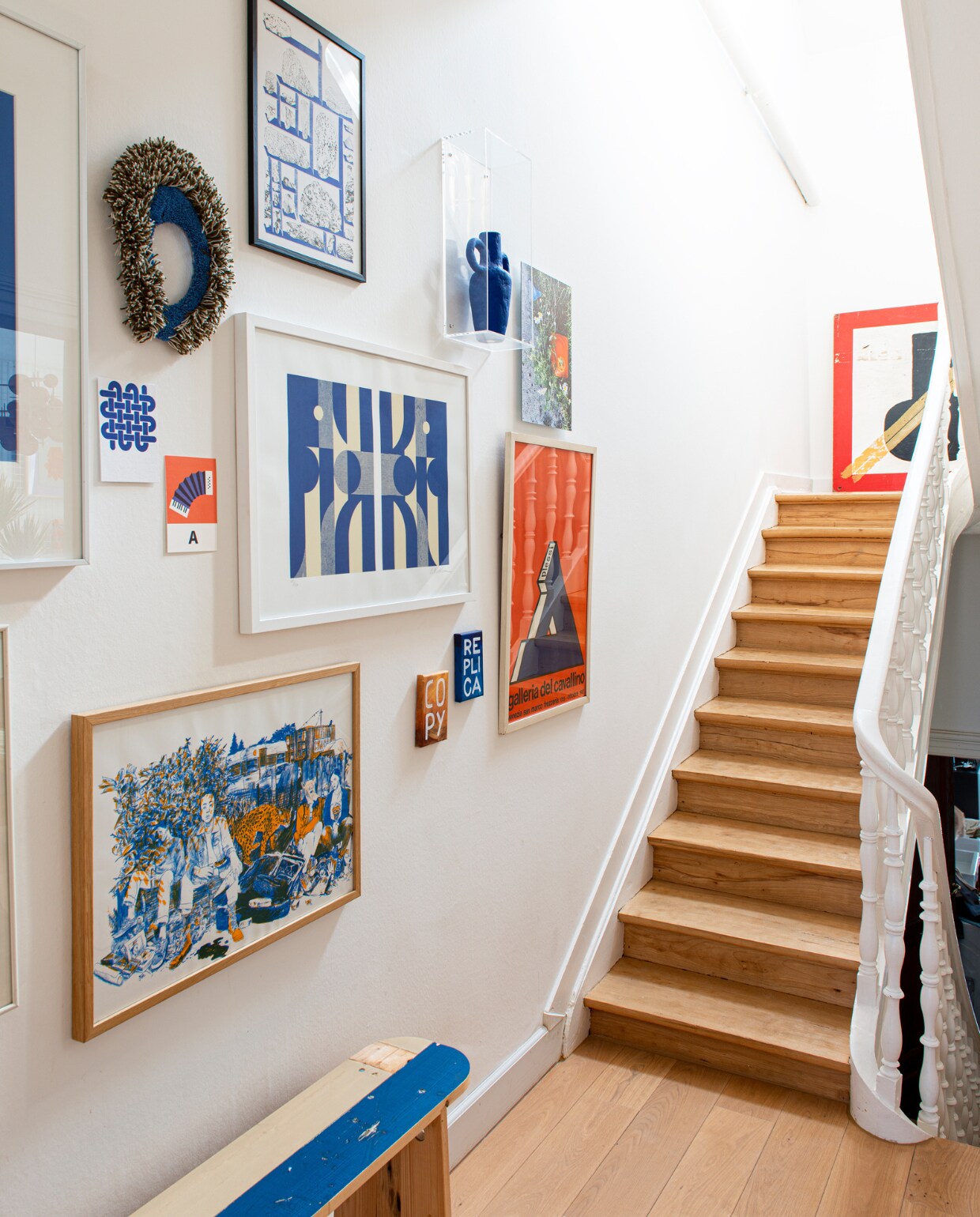 Bank Piet Hein Eek, daarboven risoprint Ward Zwart, ernaast twee kleine schilderijen van Jan Leenders, in het midden blauw-witte zeefdruk van Louis Reith, links ernaast prints Edouard Schneider, erboven rond getuft object Kihnu, helemaal boven risoprint Mélanie Corre, rechts daarvan vaas papierklei Jacqueline de la Fuente, daarnaast foto van Eline, eronder typografisch affiche tentoonstelling Plessi, bovenaan de trap een houten signaalbord voor de scheepvaart. vtwonen 02-2023 | productie De Binnenkijkers | styling Jeske Weel | fotografie Caroline Coehorst