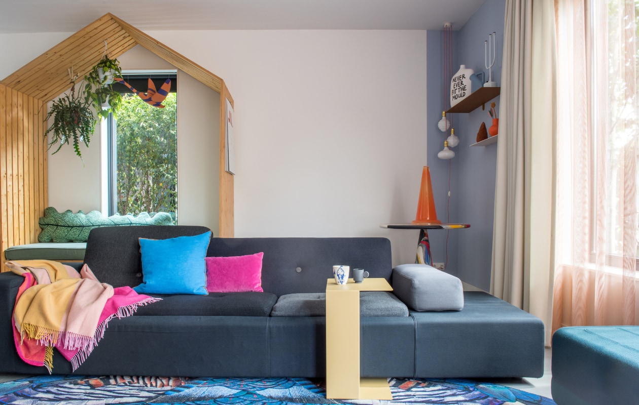 Houten huis eigen ontwerp by Maykel, bladkussen Donna Wilson, zwaan Jantien Baas. Bank Polder Sofa en plaid Hella Jongerius voor Vitra, vloerkleed Moooi Carpets, blauw kussen Hay, paars kussen Nijhof, bijzettafel Gelderland Ori, kopje links Social Label, rechts Maarten Baas, poef Normann Copenhagen. Tafel in hoek Dirk Vander Kooij, kunstwerk Save me Glass traffic cone van Bas Kosters, wandplanken Muller van Severen, vaas Irenic Mould van Birgit Verwer via Cor Unum, karaf Muuto, kandelaar Chris Kabel voor Droog Design, duif Christien Meindertsma, oranje vaas Raawii, lampen Zangra. Overgordijnen op maat gemaakt door stoffeerder, vitrage van Kvadrat-stof, kleur op muur onbekend. vtwonen 02-2022 | productie Anke Leunissen | styling Kim de Groot | fotografie Anke Leunissen