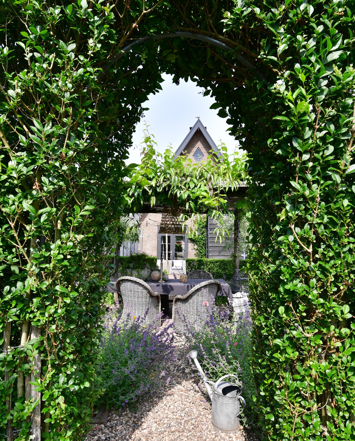 Kijkje door de ligusterboog naar de derde tuinkamer: de pluktuin. Over het dak van rietmatten groeien witte druif en Clematis. Eettafel en stoelen Avh-Outdoor tuinmeubelen. Tafelkleed en vaas Xenos. Lamp en houten bollen Loods 5. Pot TP Flowers &amp; More. Gieter rommelmarkt. vtwonen special tuin 07 | fotografie Sietske de Vries