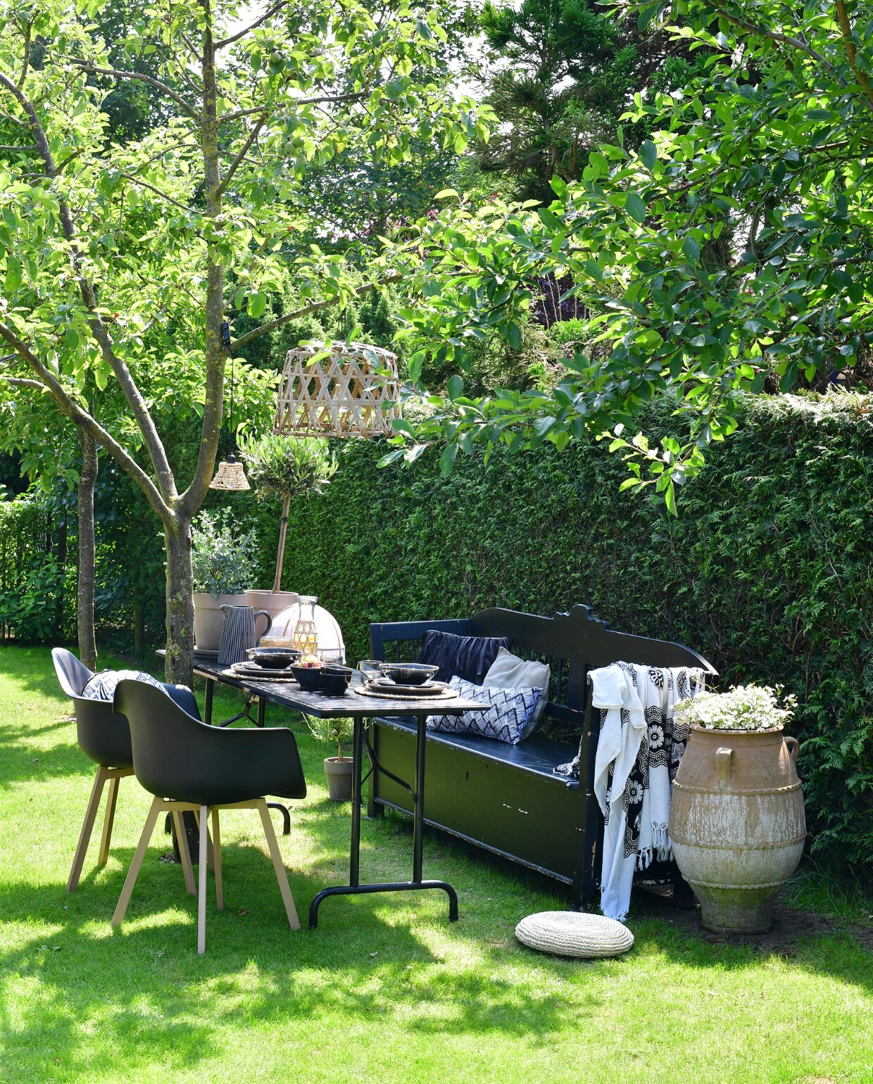 In de tweede tuinkamer is een zit- en eetplek ingericht onder de appelboom. Klepbank overgenomen van een kennis en zwart geverfd. Zwart geverfde picknicktafel gemaakt van een onderstel van de rommelmarkt en een zelfgetimmerd blad. Stoelen, lampen, plaid, kussens en kan Loods 5. Grote pot tuincentrum. Karaf, placemats, glas, bestek en  muggennet Xenos. Servies Broste Copenhagen. Potten met olijf- en eucalyptusboompje TP Flowers &amp; More. vtwonen tuinspecial tuin 7-2023 | fotografie Sietske de Vries
