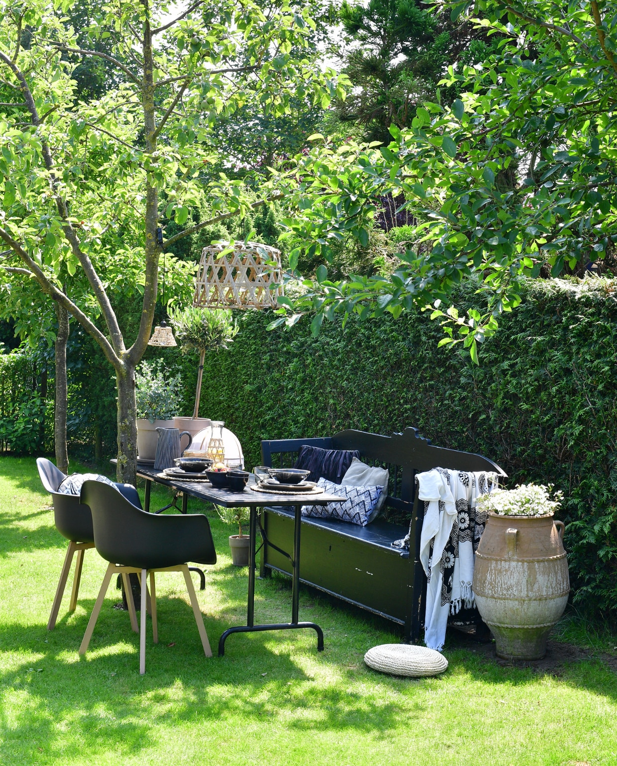 In de tweede tuinkamer is een zit- en eetplek ingericht onder de appelboom. Klepbank overgenomen van een kennis en zwart geverfd. Zwart geverfde picknicktafel gemaakt van een onderstel van de rommelmarkt en een zelfgetimmerd blad. Stoelen, lampen, plaid, kussens en kan Loods 5. Grote pot tuincentrum. Karaf, placemats, glas, bestek en  muggennet Xenos. Servies Broste Copenhagen. Potten met olijf- en eucalyptusboompje TP Flowers &amp; More. vtwonen tuinspecial tuin 7 2023 | fotografie sietske de vries