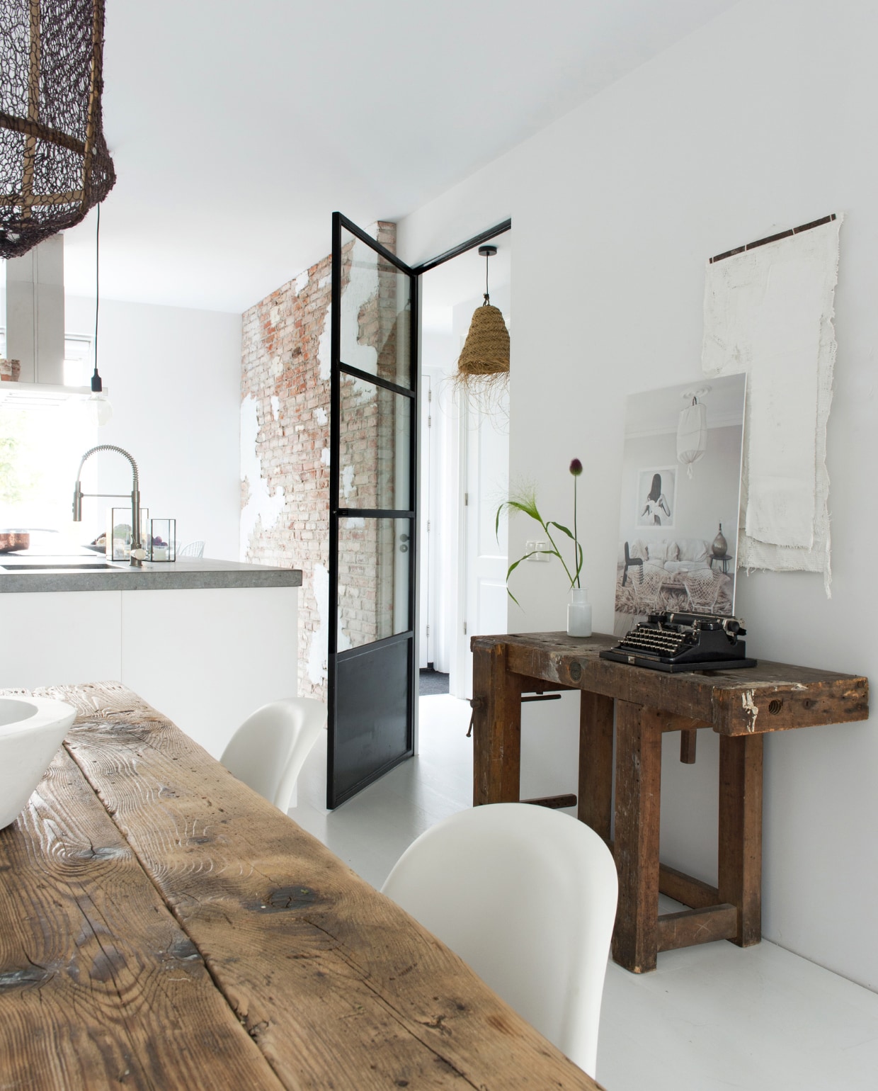De muur in de keuken bestaat uit oude bakstenen via marktplaats.nl. Stalen deur zelf ontworpen. Witte Verner Panton stoelen. Houten eettafel Barn9 Woodconcepts. Lamp Ay Illuminate loftloft.nl. Oude werkbank uit Frankrijk met een antieke typemachine. Wit kunstwerk aan de muur Neeltje Geurtsen. Foto mointerior.com. Vaasje Hema. Hanglamp gang Loods 5. vtwonen 12 binnenkijken special 2019 | Styling Wilma Custers (Features &amp; More), Rozmeen Koornneef | Fotografie Trui Heinhuis