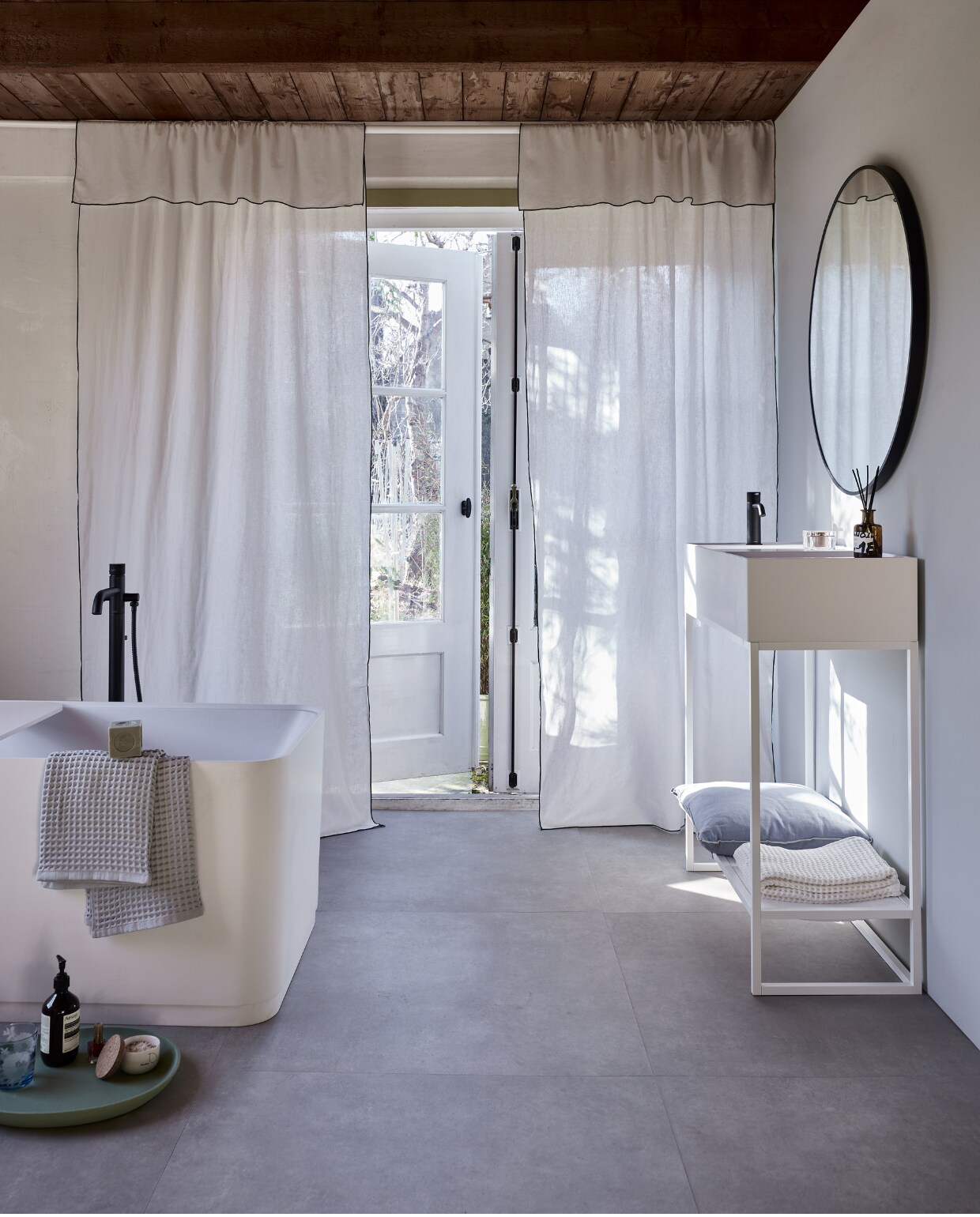 Witte gordijnen met zwarte rand, linnen en katoen, 120x250 cm (bxh), set van 2 € 59,99 (hm.com). Uitschuifbare gordijnroede Hugad, 210-385 cm (l) € 12,99,- en uitschuifbare gordijnroede Räcka, 210-385 cm (l) € 9,99,-, aan elkaar gedraaid (ikea.nl). Muurverf Tin van vtwonen, per liter € 27,99 (karwei.nl). Lichtgrijze pvc-vloer Basic XL van vtwonen, dryback, per m2 € 67,95 (vtwonen.nl). Kussen Outline in Ice Blue van Hay, katoen en linnen € 85,- (vtwonen.nl/shop). Bad Soft Edge van vtwonen, 160x73x59,5 cm (lxbxh) € 4400,-, waskom van vtwonen € 869,-, wastafelmeubel met inlegblad van No Limit € 847,-, spiegel, 90 cm (Ø) € 431,- (detremmerie.be). Wastafelmengkraan Roma € 369,-, vrijstaande badkraan Roma € 1829,- (plieger.nl). Wafelhanddoeken € 65,- (hay.nl). Groen dienblad, set van 3 € 175,-(polspotten.nl). vtwonen 05-2023 | styling Marianne Luning | fotografie Alexander van Berge