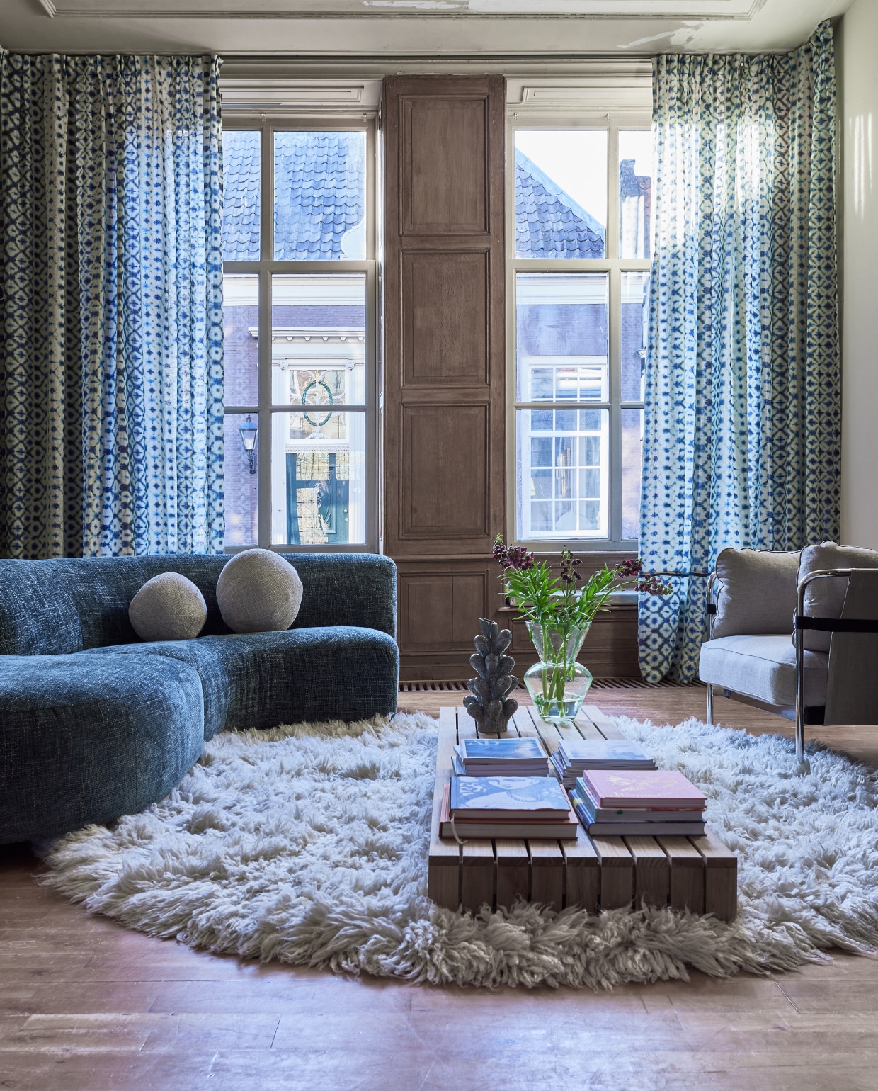 Blauwe linnen stof Karaoshi van Designers Guild, kamerhoog 295 cm, per meter € 200,-, donkerblauwe polyester stof voor band op gordijn Chaviere Indigo van Designers Guild, kamerhoog 300 cm, per meter € 112,-, gestreepte linnen stof voor matrashoes Brera Strada van Designers Guild, 140 cm (b), per meter € 132,- (wva.nl). Aluminium gordijnrail met afgeronde zijkant van 30 cm, verkrijgbaar in alle Ral-kleuren, per meter € 65,- (xxldesignroeden.nl). Modulebank A-Round, 167x167x74 cm (bxdxh) € 1995,-, zij-element Poef A-round, 100x100x46 cm (bxdxh) € 645,-, polyester kussen Ball S € 95,-, polyester kussen Ball L € 129,-, glazen vaas Fat Neck, 25x35 cm (Øxh) € 159,-(polspotten.nl). Vloerkleed Fluffy, wol en katoen, 250 cm (Ø) € 1575,- (hkliving.nl). Fauteuil Can, staal en canvas, 97x89x82 cm (bxdxh) € 2059,-(hay.nl). Teakhouten salontafel Lounge van HKliving, 60x200x15 cm (bxdxh) € 995,-, keramieken vaas Innes van Bloomingville, 18,5x36,5 cm (Øxh) € 99,90 (vtwonen.nl/shop). Boeken privébezit.
