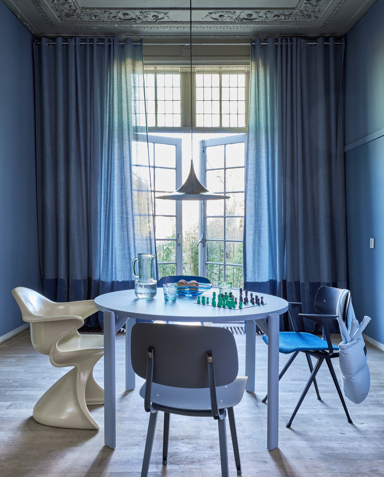 Blauwe gordijnstof Tick-Tock Crushed Reboot van Chivasso, linnen en polyester, kamerhoog 300 cm, per meter € 147,50, lichtblauwe fluwelen stof Sensual Velvet van Chivasso, polyester en katoen,140 cm breed, per meter € 105,- (jab.de). Rvs gordijnroede inclusief steunen en eindknoppen, per meter vanaf € 115,- (xxldesignroeden.nl). Krijtverf Classico in Blue Stone, per liter € 55,75 (pure-original.com). Houten tafel Rey, 128,5x74,5 cm (Øxh) € 2049,-, blauwe eetkamerstoelen Revolt € 385,-, Result € 509,- en Rey € 549,-, kartonnen schaakspel van Hay € 55,-  (hay.nl). Hanglamp € 490,-(goeds.nl). Roomgele plastic stoel Sedia Sabbia € 895,- (hkliving.nl). Tas € 99,95 (madsnorgaard.com). Gekleurde drinkglazen, set van 6 € 119,-, glazen schaal Eye € 125,-, glazen waterkan € 62,50 (polspotten.com). vtwonen 05-2023 | styling Marianne Luning | fotografie Alexander van Berge