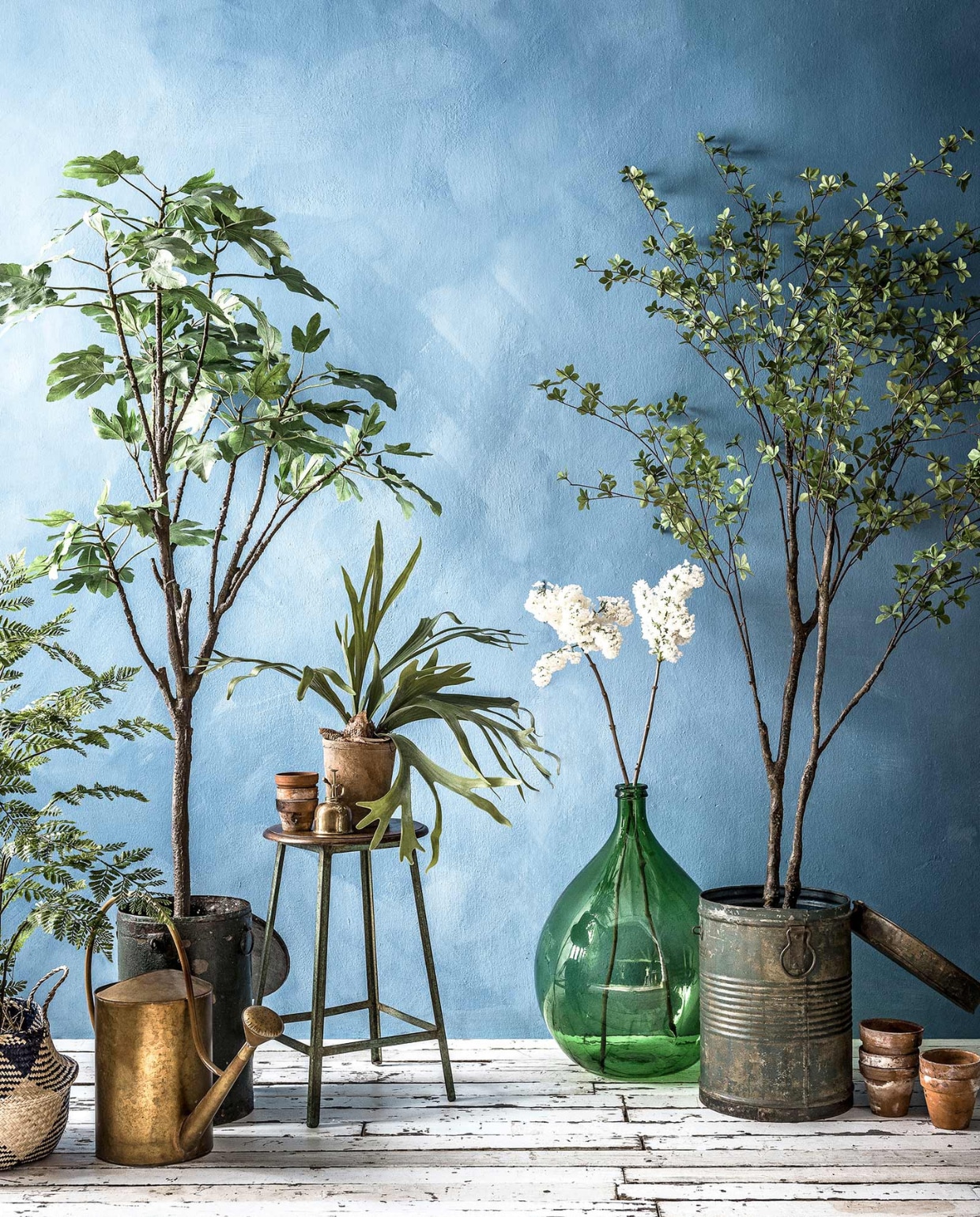 Muurverf in kleur Periwinkle van Bauwerk Color (). Gevlochten mand met blauwe vlakken (). Kleine kunstplanten in pot, grote kunstplanten en -boompjes, groene metalen ton met deksel, houten kruk met metalen onderstel (). Messing gieter (). Oude terracotta bloempotten, witte seringen () Messing plantenspuit (). Groene glazen flesvaas () vtwonen 2017-08 | fotografie Sjoerd Eickmans | styling Danielle Verheul