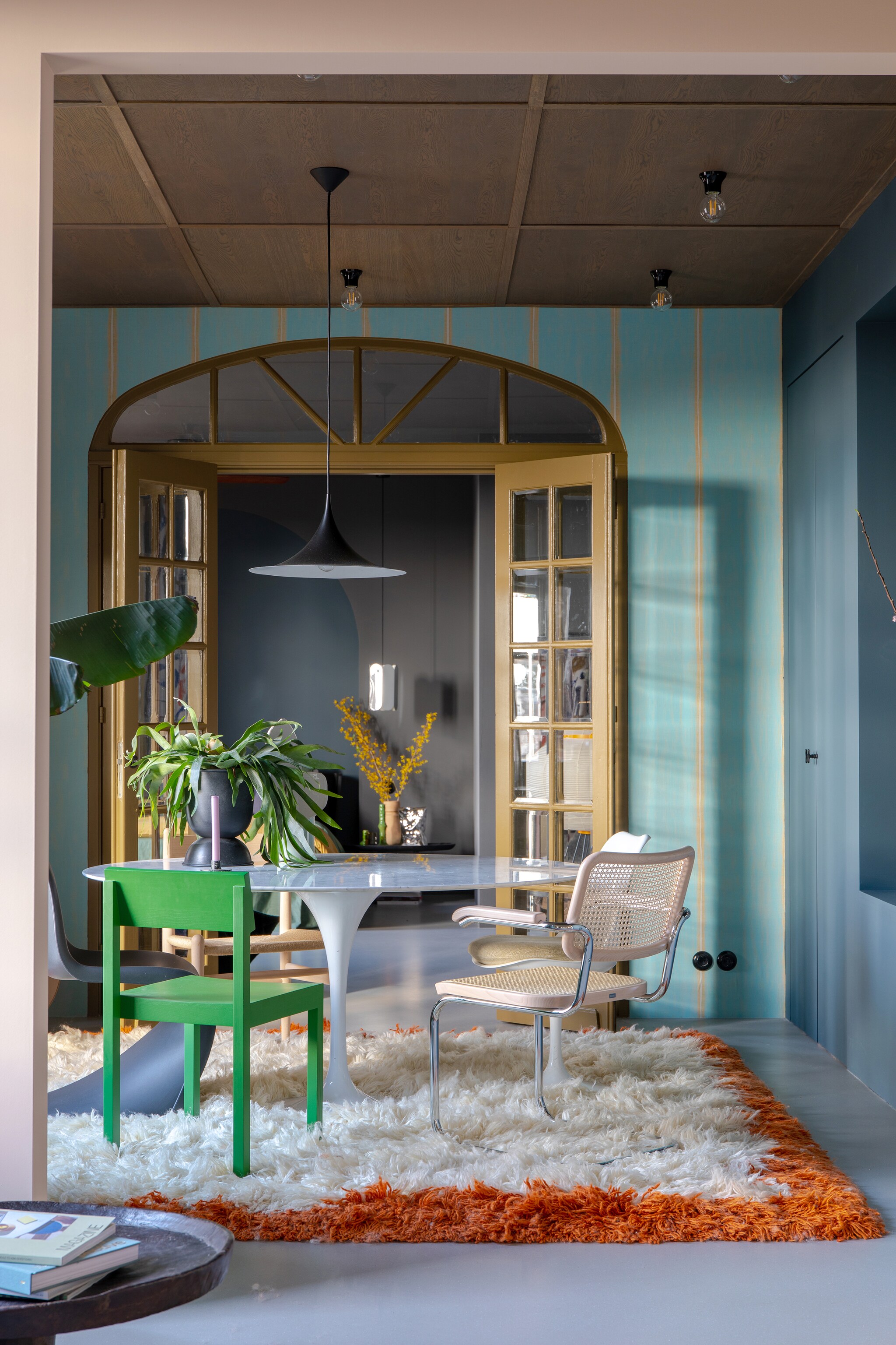 Muurverf 85% Graniet en Puur Marmer van Flexa, behang via Selected Wallpaper. Groene eetkamerstoel Valerie Objects, rotan stoel Thonet, witte stoel Tulip van Eero Saarinen, houten stoel Carl Hansen, zwarte stoel Panton van Vitra, vloerkleed HKliving. Plafond op maat gemaakt door Woodever, hanglamp Gubi, bakelieten plafondlampen Fonq, zilveren hanglamp Hay. Groene kandelaar en chipszak-vaas HKliving, aardewerken vaas MaisonNL, eettafel en en-suitedeuren vintage. Overig onbekend vtwonen 05-2023 | Styling &amp; fotografie Theo-Bert Pot &amp; Jelle van de Schoor