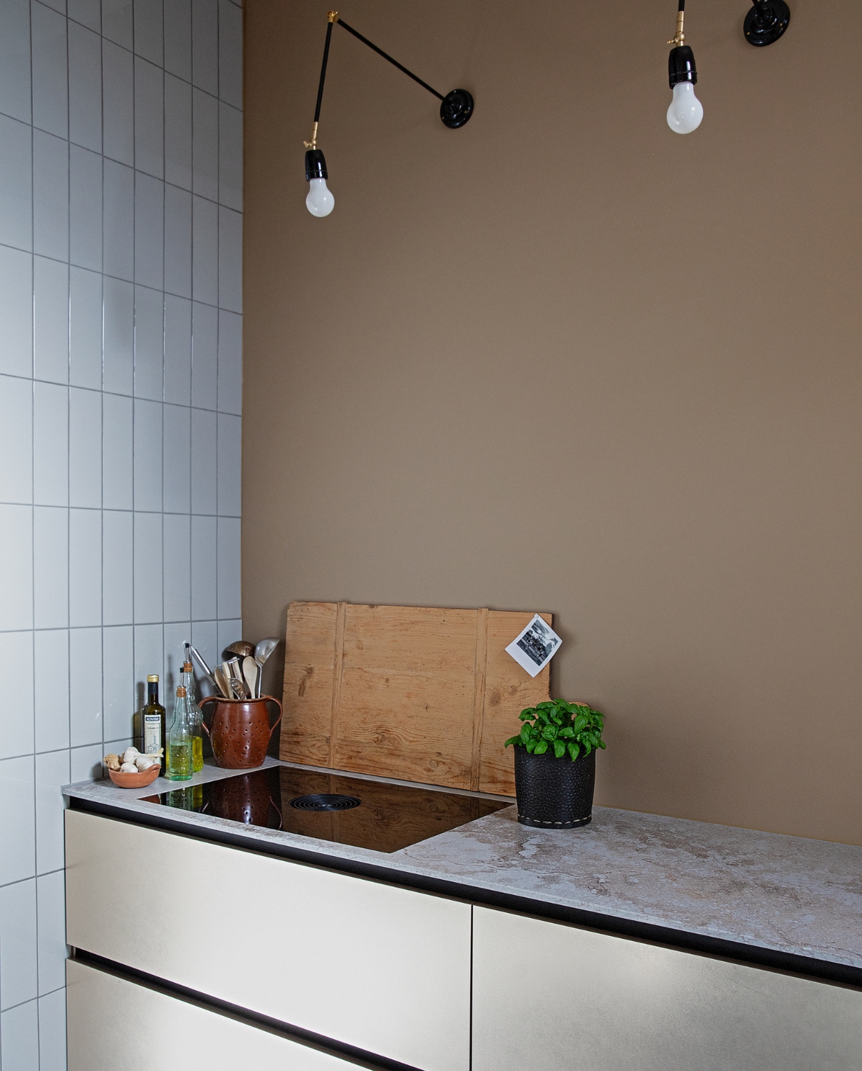 Tegels Praxis, muurverf Grijsbeige Ral 1019 Flexa, greeploze keuken met goudkleurige fronten en composiet werkblad Brugman Keukens, kookplaat Bora, wandlampen Zangra, houten plank Carlotte Antiek en Brocante, rubberen mand voor basilicum Sissy-Boy, pot voor lepels uit Duitsland, schaal voor knoflook Bij Lokaal, grote olijfoliefles Van-Verre, kleine olijfoliefles en limonadefles uit Italië. vtwonen 04-2021 | productie en styling Ilona Jongepier | fotografie Dana van Leeuwen