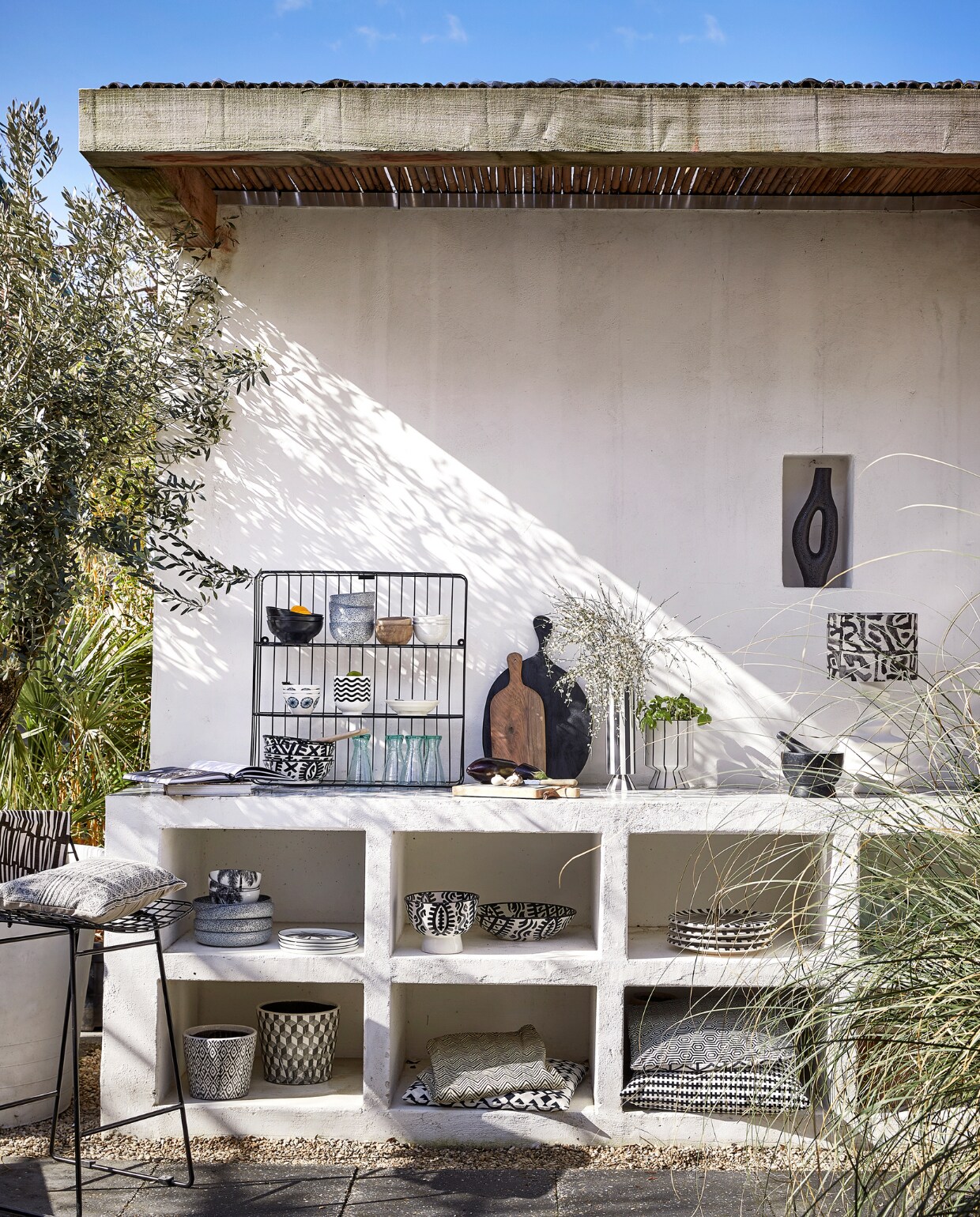 Metalen wandrek, 60x60 cm € 44,95, kussen op kruk € 22,50 (mrsbloom.nl). Metalen barkruk Wire, 47x42x82 cm(bxdxh) € 219,- (hkliving.com). In het rek: zwarte kommen Bold &amp; Basic, per 4 € 25,95, kommen Gradient, per 4 € 34,-, witte kommen Kyoto € 6,-(hkliving.nl), houten kommen € 12,95 (householdhardware.nl), witte gebloemde kom € 1,99, bloempot met golven € 3,99 (ikea.nl), schaal, per 4 € 33,56 (hkliving.com), Marokkaanse kommen Fex, per stuk € 55,50, houten lepel € 5,-, glazen Diabolo uit Marokko € 6,- (householdhardware.nl). Op het keukenblok: walnoothouten plank € 37,50 (householdhardware.nl), zwarte houten serveerplank € 42,95, leistenen snijplank € 24,95, gestreepte vaas Toppu van Oyoy € 69,95, pot op voet € 19,95 (livvlifestyle.nl). Spekstenen vijzel € 34,95 (dille-kamille.nl). Lampenkap Adir € 69,90 (ateliersem.nl). Organische vaas in nis € 51,95 (ptmd.nl). In de vakken: diepe borden Gradient, per stuk € 35,-, kommen met palmboom € 7,95, ontbijtbord met palmboom € 9,95 (hkliving.com), saladeschaal Fez uit Marokko € 129,-, grote bowl Fez € 55,-, borden Fez Assiette € 79,95 (householdhardware.nl), bloempot met ikatprint € 28,95, pot met geometrische print € 51,95, stoelkussen onder met grafische print € 45,95 (ptmd.nl). Tafellinnen € 64,95 (mrsbloom.nl). Kussen boven € 45,95 (ptmd.nl), kussen Stockholm € 19,99 (ikea.nl). vtwonen 06-2023 | styling Kim Timmerman | fotografie Peggy Janssen