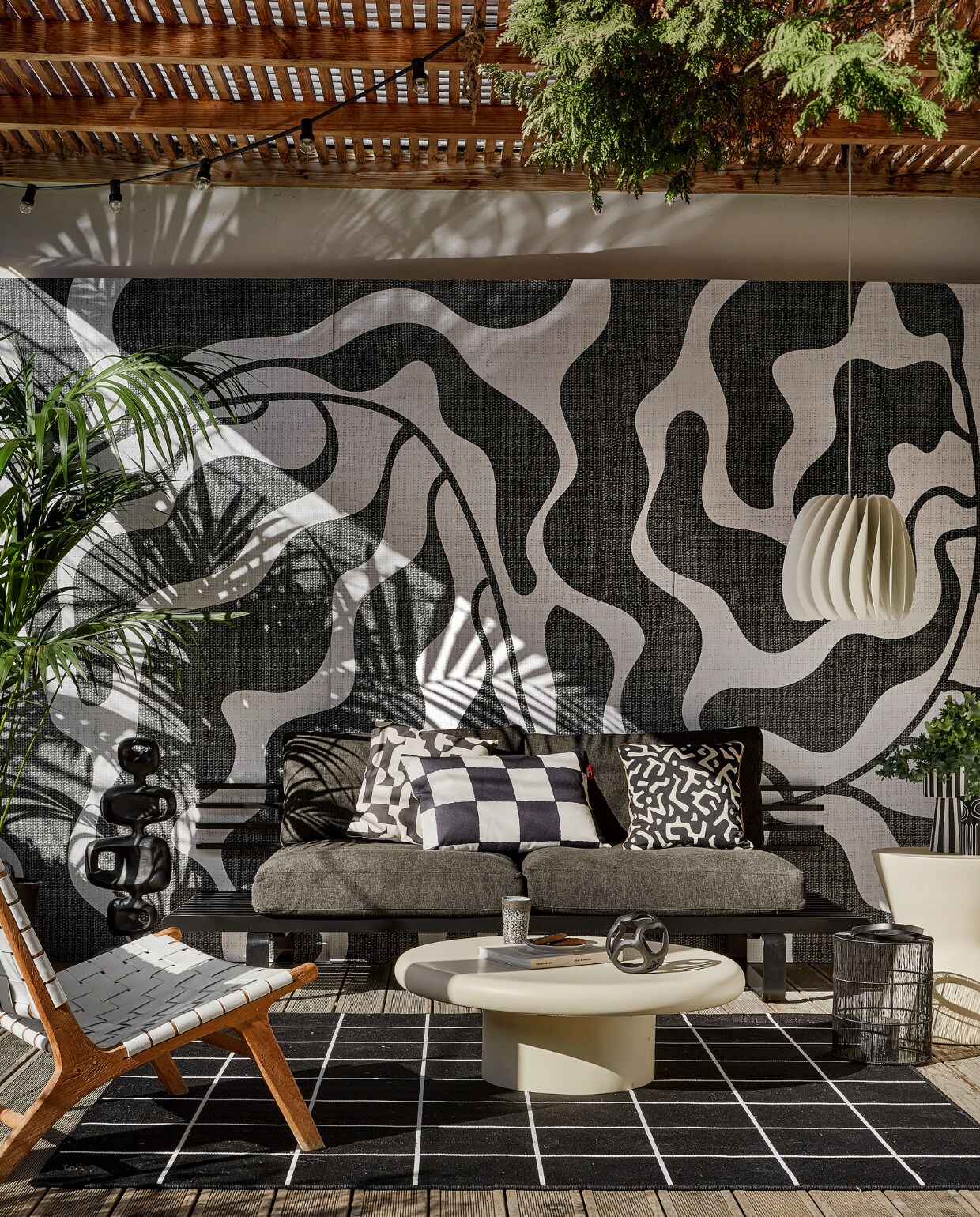 Behang River black &amp; white van Wrap the Wall, per m2 € 150,-, minimale afname 4 m2 (behangfabriek.nl). Zwarte aluminium buitenbank € 1339,80, antraciet tuinkussens, per set € 675,75, zwarte aluminium sculptuur € 295,-, mok 70s, per 2 € 19,99 (hkliving.com). Buitenvloerkleed Svallerup, 200x200 cm € 79,95, hanglamp Skymningen € 79,95 (ikea.nl). Ivoorkleurige polyester bijzettafel, 85x30 cm (Øxh) € 599,-, bijzettafel Pot-Table in Ivory-Slim € 395,- (householdhardware.nl). Zwart-wit kussen Cutro € 10,- (kwantum.nl). Zwart-wit geblokt kussen King Pillow € 55,- (fatboy.com). Kussen Nara € 59,90 (ateliersem.nl). Zwart-wit gestreepte pot van Oyoy € 69,95, aardewerken vaas Troy van Bloomingville € 12,95 (livvlifestyle.nl). Zwart aluminium bolvormig ornament € 31,95, lantaarn € 96,95 (ptmd.nl). Fauteuil met gevlochten zitting privébezit, soortgelijke bij Kave Home (kavehome.com). vtwonen 06-2023 | styling Kim Timmerman | fotografie Peggy Janssen
