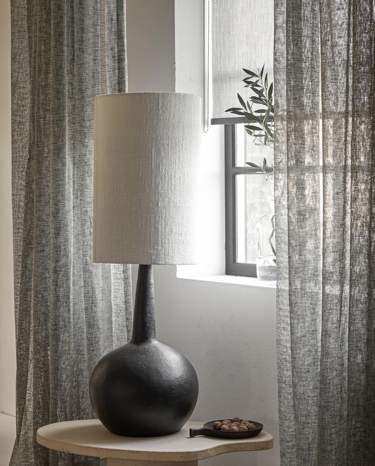 Inbetween gordijn op maat met enkele plooi in stof Weave (2712) kleur Brush, vanaf vanaf € 32,99 per strekkende meter. Lichtdoorlatend rolgordijn op maat in kleur antraciet (18015), vanaf € 128,19. M.m.v.: rawmaterials.eu (tafellamp Loulou XL), bogard.eu (salontafel Palet 80x50 cm in kleur Stone, afgewerkt in beton op MDF), vtwonen.nl/shop (schaal met handvat van zwart mangohout). vtwonen 06-2023 | productie Tanja Saarberg | styling Marit Saladini Platjouw | fotografie Peggy Janssen