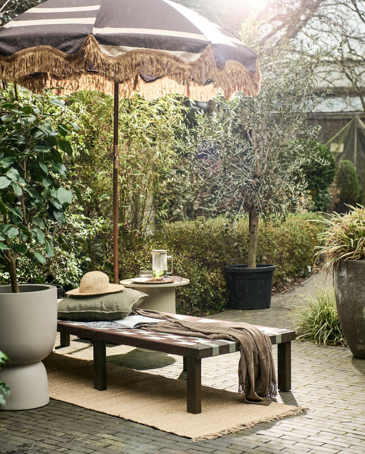 Grenenhouten frame daybed, 200x70x31,5-77 cm (lxbxh) € 67,99 (vidaxl.nl). Parasol van HKliving € 369,- (bijenkorf.nl). Grijze bloempot van Elho € 145,-, bijzettafel van HKliving € 249,- (vtwonen.nl/shop). Plaid € 24,99, kussen € 12,99, juten vloerkleed € 42,99 (hmhome.nl). Glazen karaf € 17,99, houten dienblad € 17,99, glas € 5,99 (zarahome.com). Overig privébezit. vtwonen 06-2023 | styling en uitvoering Sanne Ketelaar | fotografie Sjoerd Eickmans