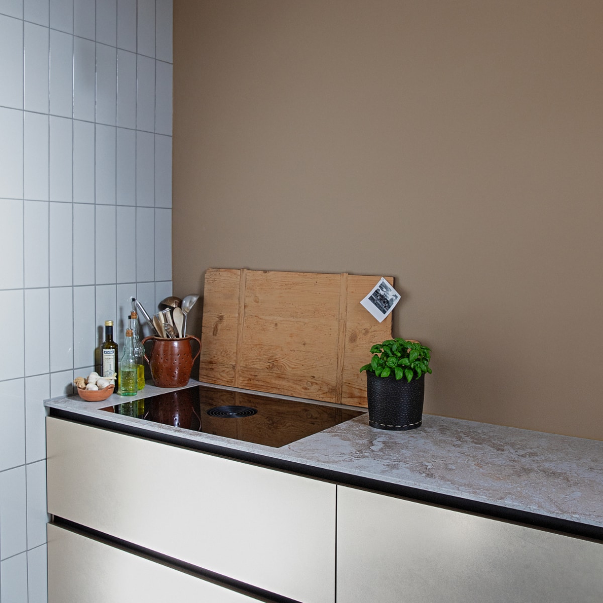 Tegels Praxis, muurverf Grijsbeige Ral 1019 Flexa, greeploze keuken met goudkleurige fronten en composiet werkblad Brugman Keukens, kookplaat Bora, wandlampen Zangra, houten plank Carlotte Antiek en Brocante, rubberen mand voor basilicum Sissy-Boy, pot voor lepels uit Duitsland, schaal voor knoflook Bij Lokaal, grote olijfoliefles Van-Verre, kleine olijfoliefles en limonadefles uit Italië. vtwonen 04-2021 | productie en styling Ilona Jongepier | fotografie Dana van Leeuwen
