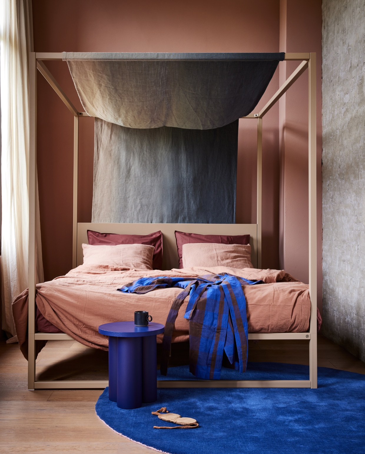 Muurverf krijtverf Waaltje (lauthentique.nl). Zandkleurig bedframe met hoofdbord 02 in Clay, gerecycled staal, 200x180x210 cm (lxbxh), matras (robuust.com). Linnen stof Padua Degrade van Designers Guild, van off black tot zand, 140 cm (b) (wva.nl). Roestkleurig linnen dekbedovertrek, donkerrood katoenen dekbedovertrek  (suite702.com). Kruk Oly van noo.ma, aluminium en hout, badjas Field van Ferm Living, katoen en linnen (vtwonen.nl/shop). Blauw vloerkleed Outline, wol en katoen, 200 cm (Ø) (polspotten.nl). Mok van Nordal, set van 4 (deens.nl). Slaapmasker (nordal.eu). vtwonen 08-2023 | styling Marianne Luning | fotografie Alexander van berge