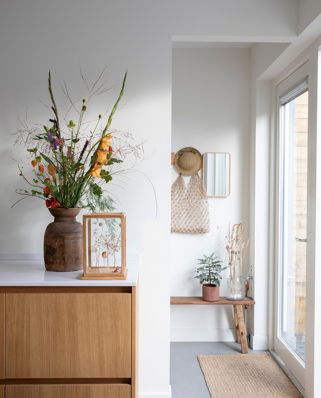 Kast onderdeel van de keuken door Keukenontwerpers Woerden. Vaas gevonden op een brocantemarkt. Vierkante stolp van Leen Bakker gevuld met droogbloemen. Wandspiegel Ikea. Hoed Zara. Gehaakte tas Daybydee Lifestyle. Bank Loods 5. Deurmat Home Delight. Herkomst pot en vaas onbekend. vtwonen 08-2021 | fotografie Macha Gutlich