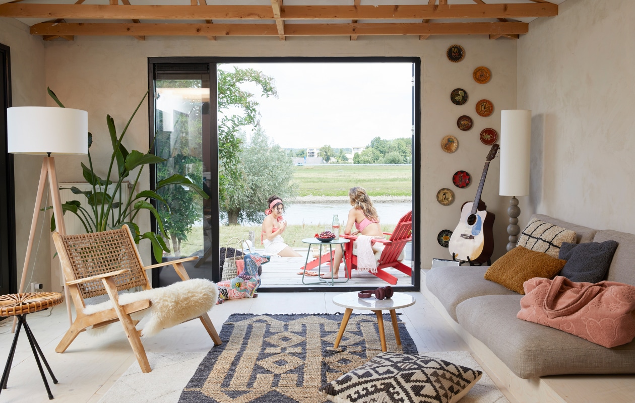 Fauteuil Grignoon van Kave Home, lampen gemaakt voor OT Design, rieten bijzettafel erfstuk. Kunstwerk teckel gemaakt door Lisa. Vloerkussen gemaakt van stof van Chivasso. Handgeschilderde wandschotels meegenomen uit Sumatra, grijze kussens oude bank. Mand uit Afrika, salontafel Zuiver, kussens en tafel buiten Karwei, strandtas meegenomen van Ibiza, rode Adirondack-stoel vidaXL. Underlayment vloer gelakt. Overig onbekend. vtwonen 08-2023 | styling Linda van der Ham | fotografie Simone de Geus