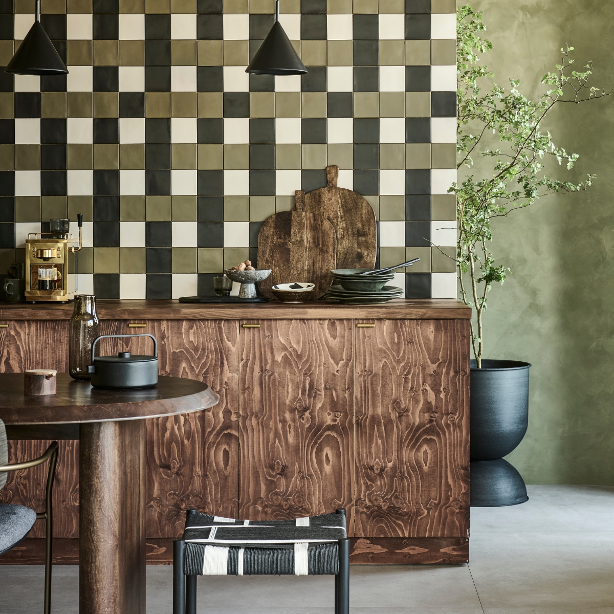 Op de wand mix van tegels Chop Kaki, Chop Nero en Chop Bone, 10x10 cm, per m²  € 79,90. Op de vloer tegels Raw Grey Mat, 120x120 cm, per m² € 92,50.M.m.v. pure-original.com (muurverf Olive Drab), basiclabel.nl (mangohouten tafel Ellips van vtwonen, kruk Jessy van Woood), hm.com (mangohouten snijplanken, marmeren etagère, zwarte snijplank, karaf, mangohouten lepels), debijenkorf.nl (schaaltjes van HKliving), zarahome.com (borden, houten suikerpot, mokken), vtwonen.nl/shop (bloempot van Ferm Living, stoel van Kave Home, hanglampen van HKliving, theepot van Serax), sjostrandcoffee.com (espressomachine). vtwonen 09-2023 | productie Tanja Saarberg | styling Sanne Ketelaar | fotografie Sjoerd Eickmans